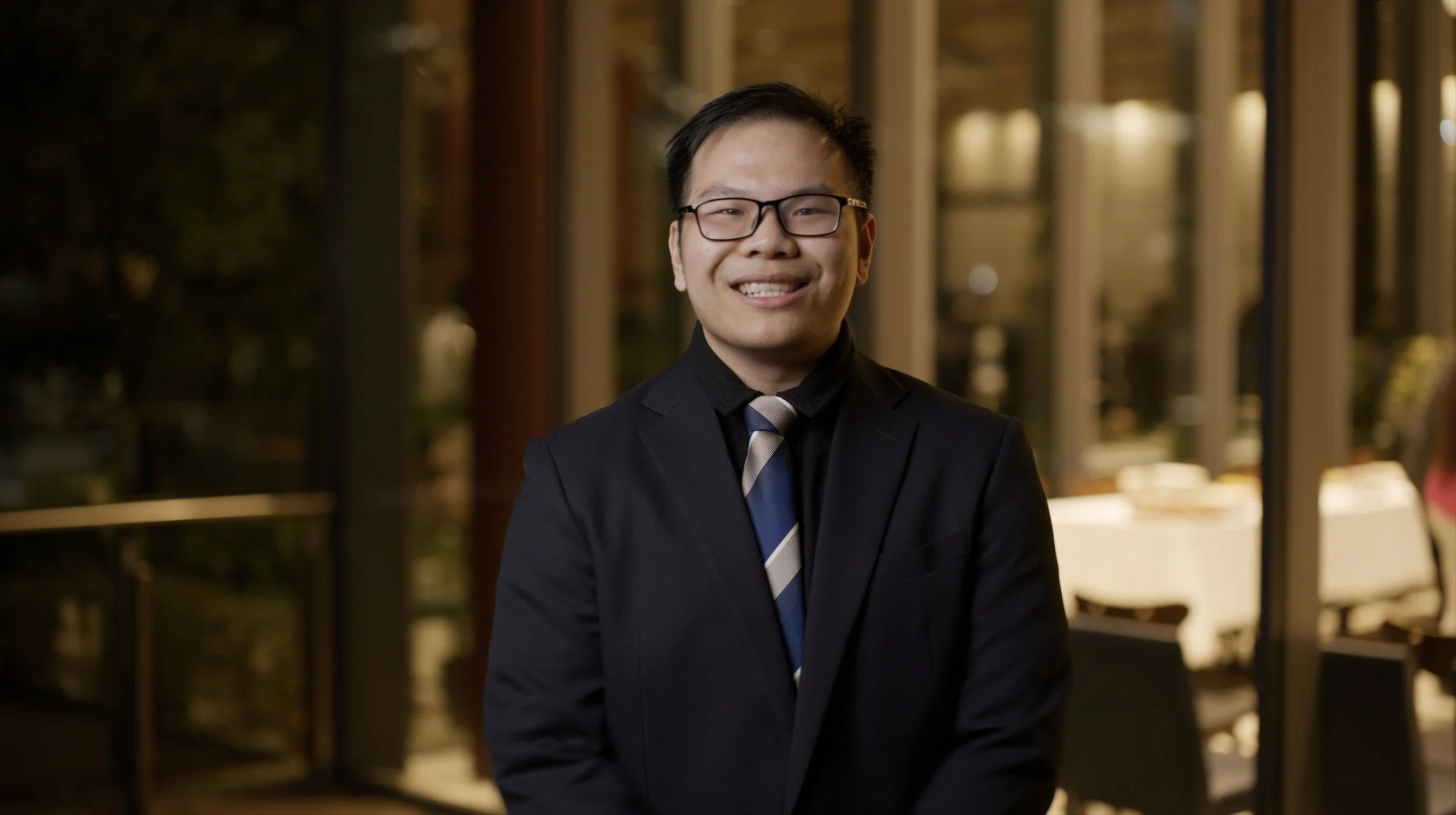 A smiling young man in a black suit, striped tie, and glasses standing inside a well-lit restaurant or café with large windows and tables in the background.