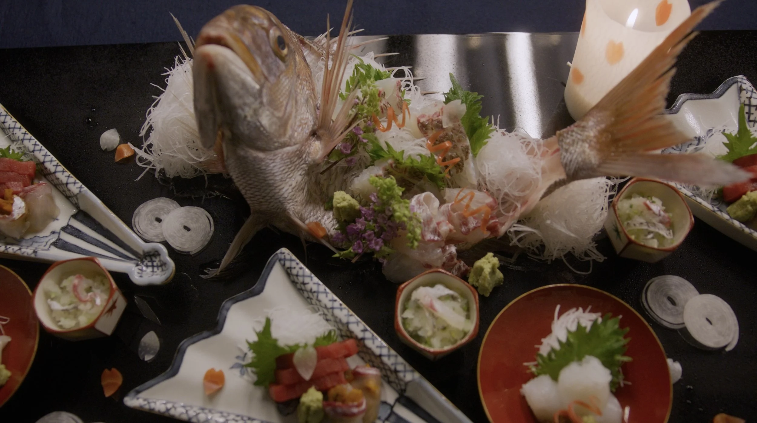 A whole fish displayed on a plate with garnishes and vegetables, accompanied by small bowls of dipping sauces or condiments, presented in a traditional Japanese style.