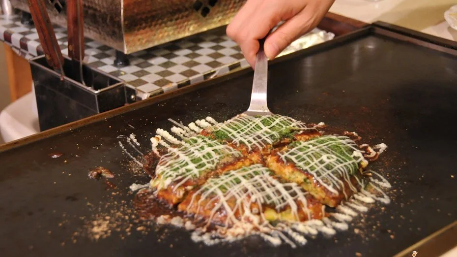 A hand holds a spatula over a cooked Japanese dish called okonomiyaki on a hot griddle, topped with sauces and garnished with a white drizzle.
