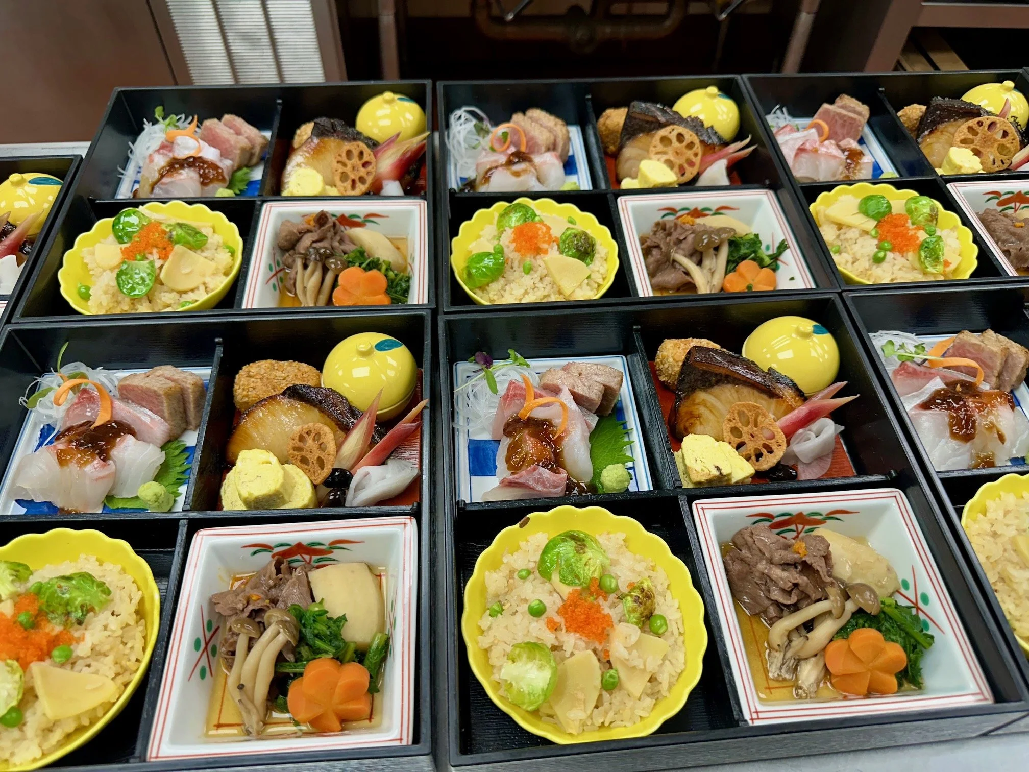 Multiple traditional Japanese bento boxes filled with rice, grilled fish, vegetables, and garnishes, arranged in black containers.