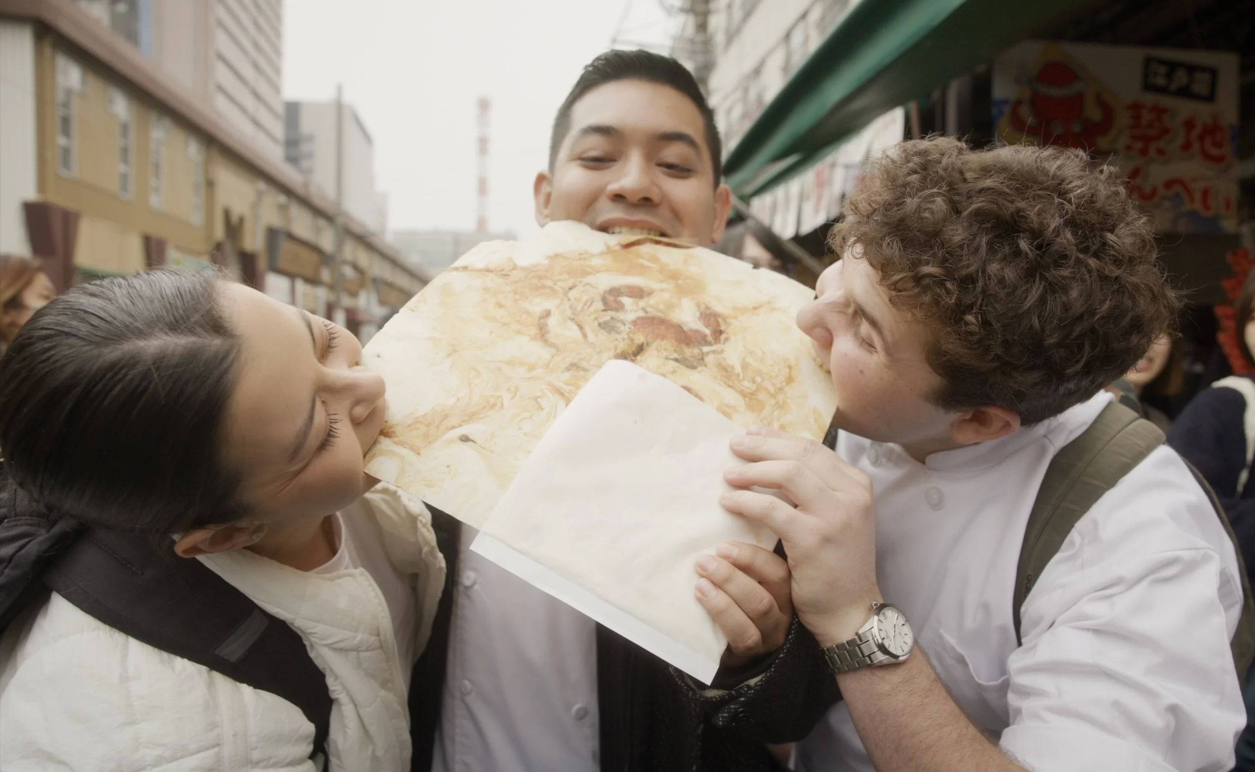 Two children and a young man sharing a large, marbled flatbread or pizza on a busy outdoor street market.