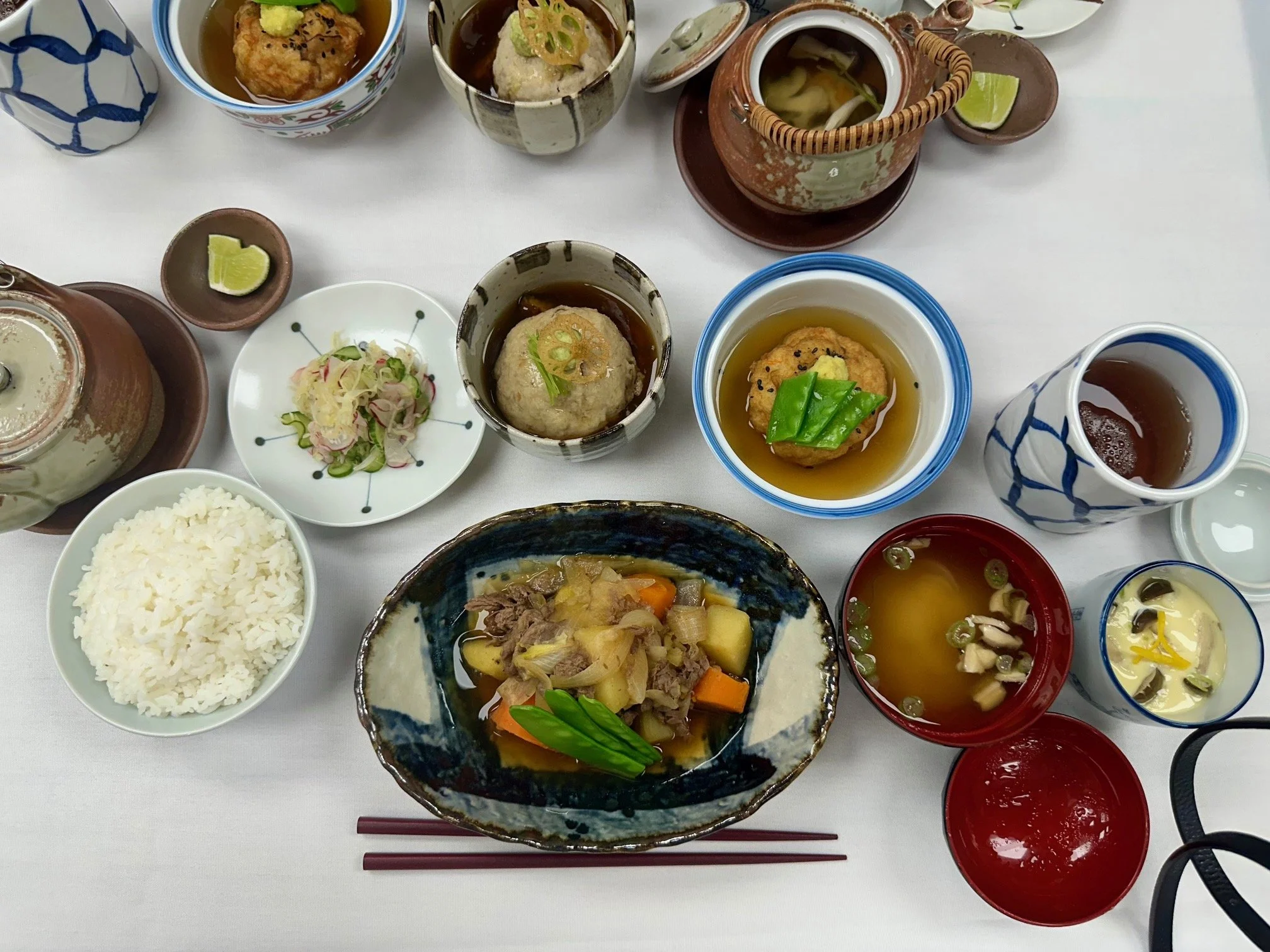 A variety of Japanese dishes served on a table, including rice, vegetable stew, meatballs in sauce, clear soup with vegetables, and small side dishes in colorful bowls.