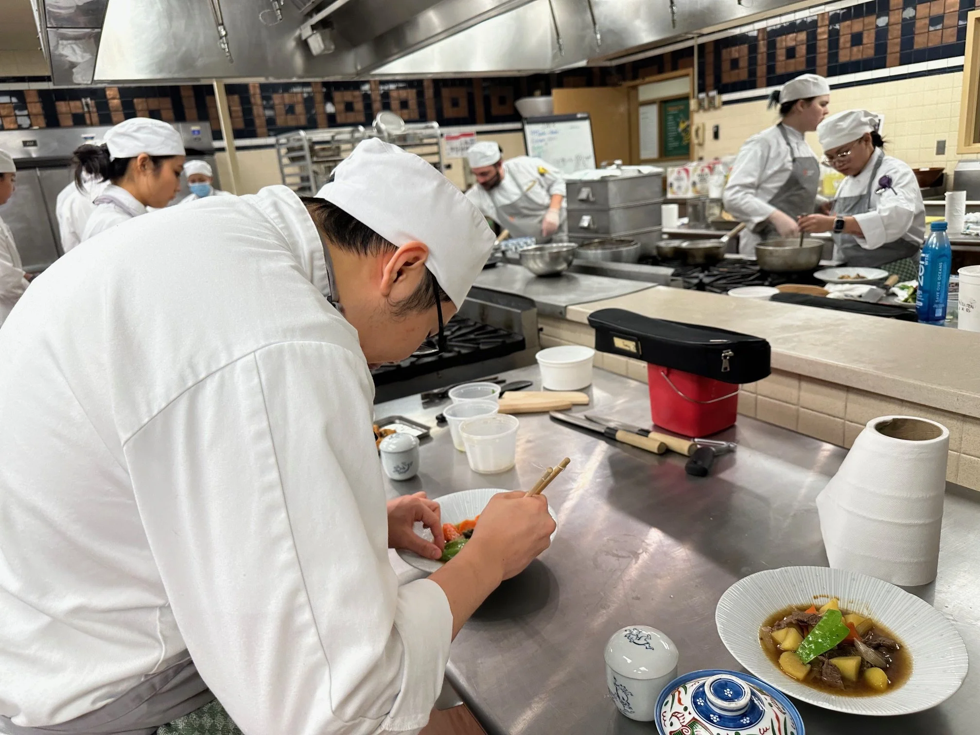People working in a professional kitchen, including a chef preparing a dish in the foreground, with several other chefs cooking and preparing food in the background.