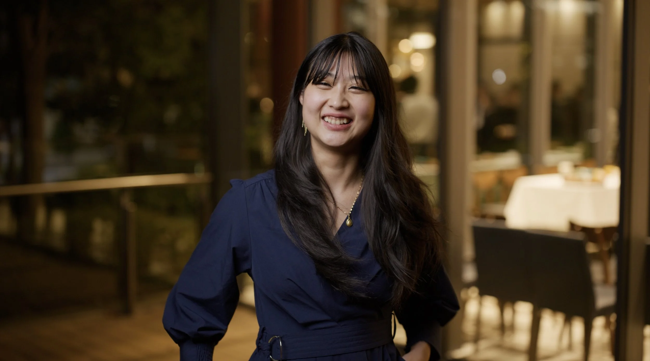 A woman with long dark hair smiling in a dimly lit outdoor restaurant or patio at night, wearing a navy blue dress with long sleeves and gold jewelry.