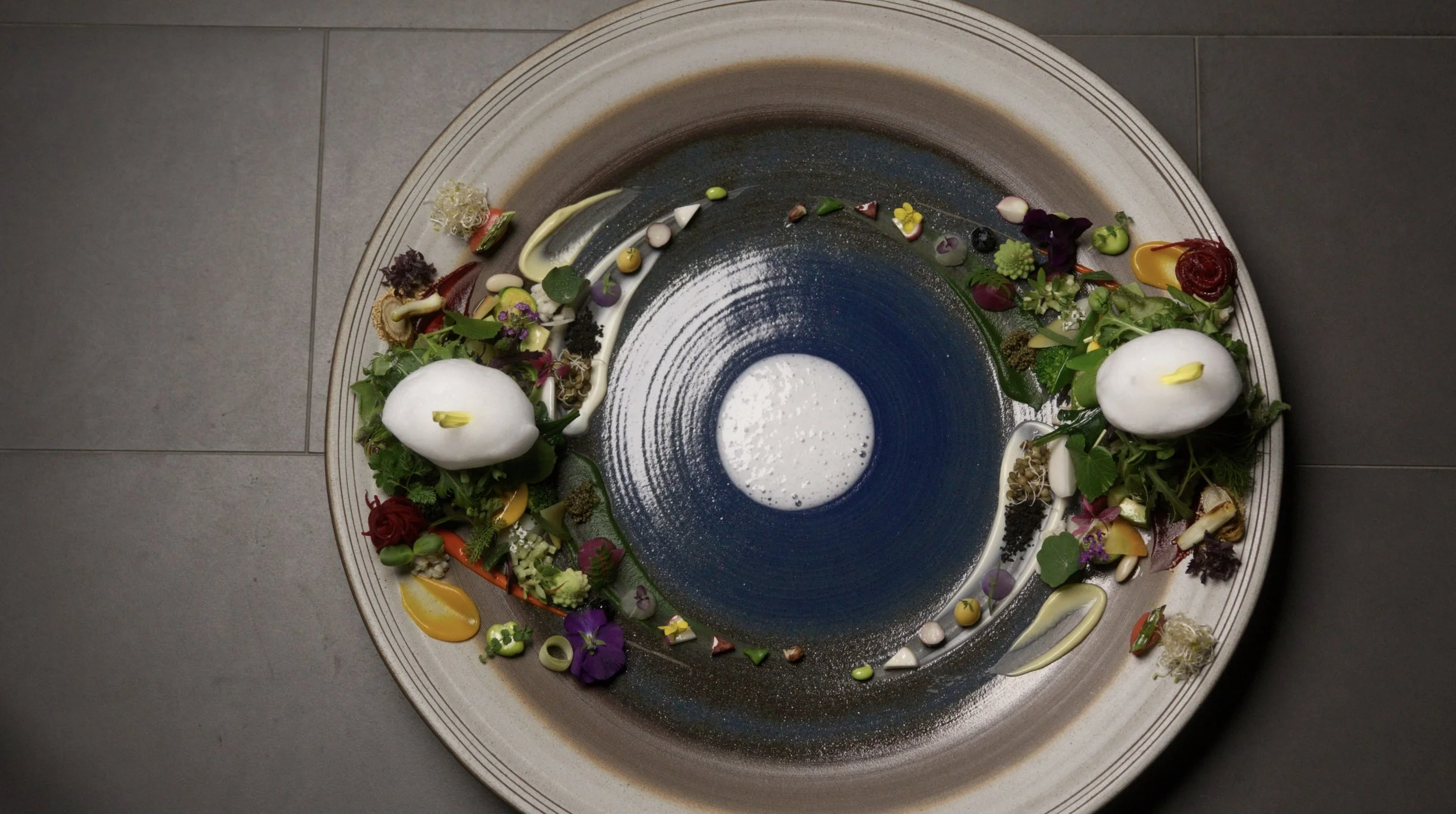 A modern, artistic seafood dish served on a large, round plate with a concentric color pattern. The plate features two egg-shaped elements, various colorful edible flowers, sauces, and garnishes arranged decoratively around the center, creating an ab