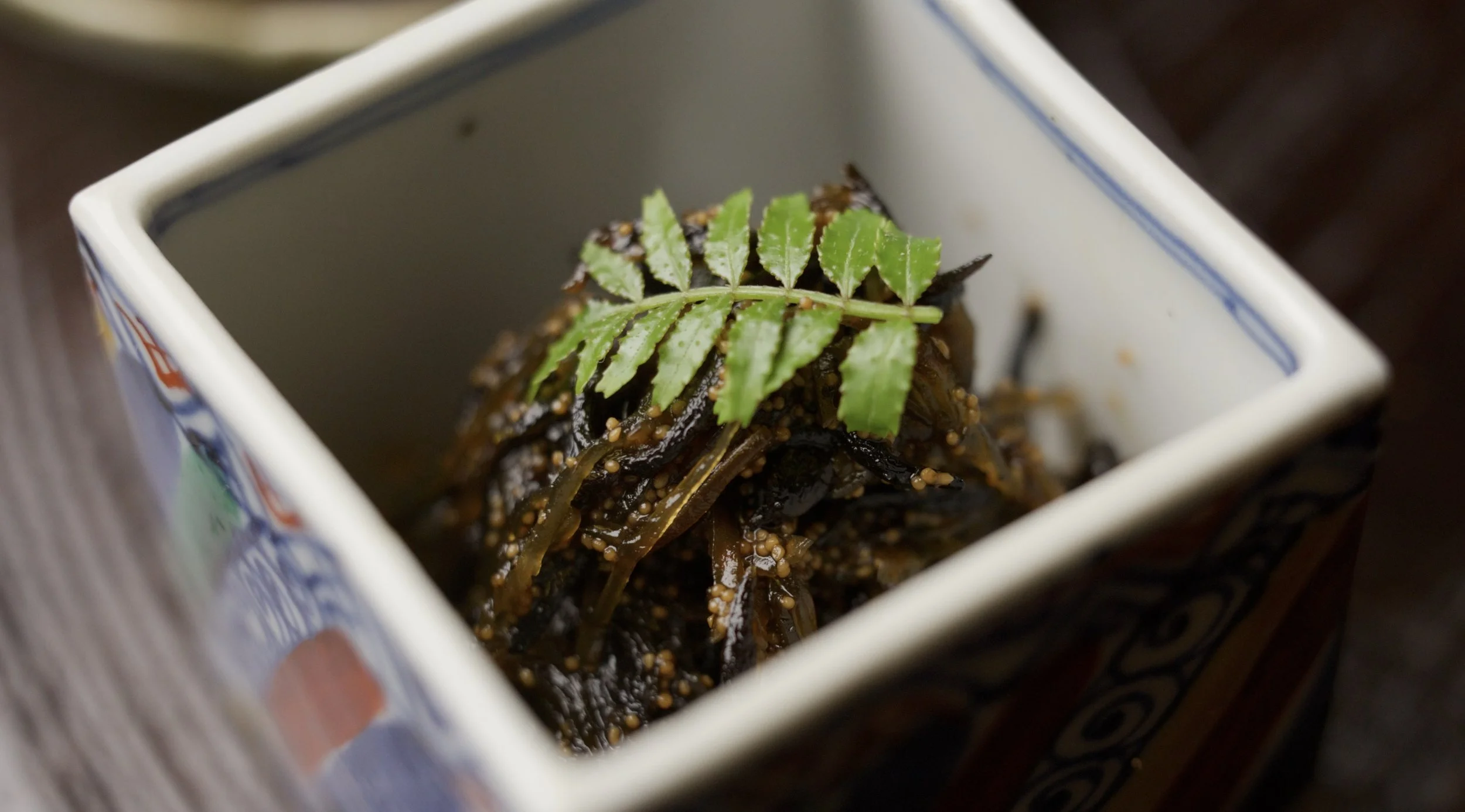 A small square dish with seaweed salad garnished with a sprig of green herb.
