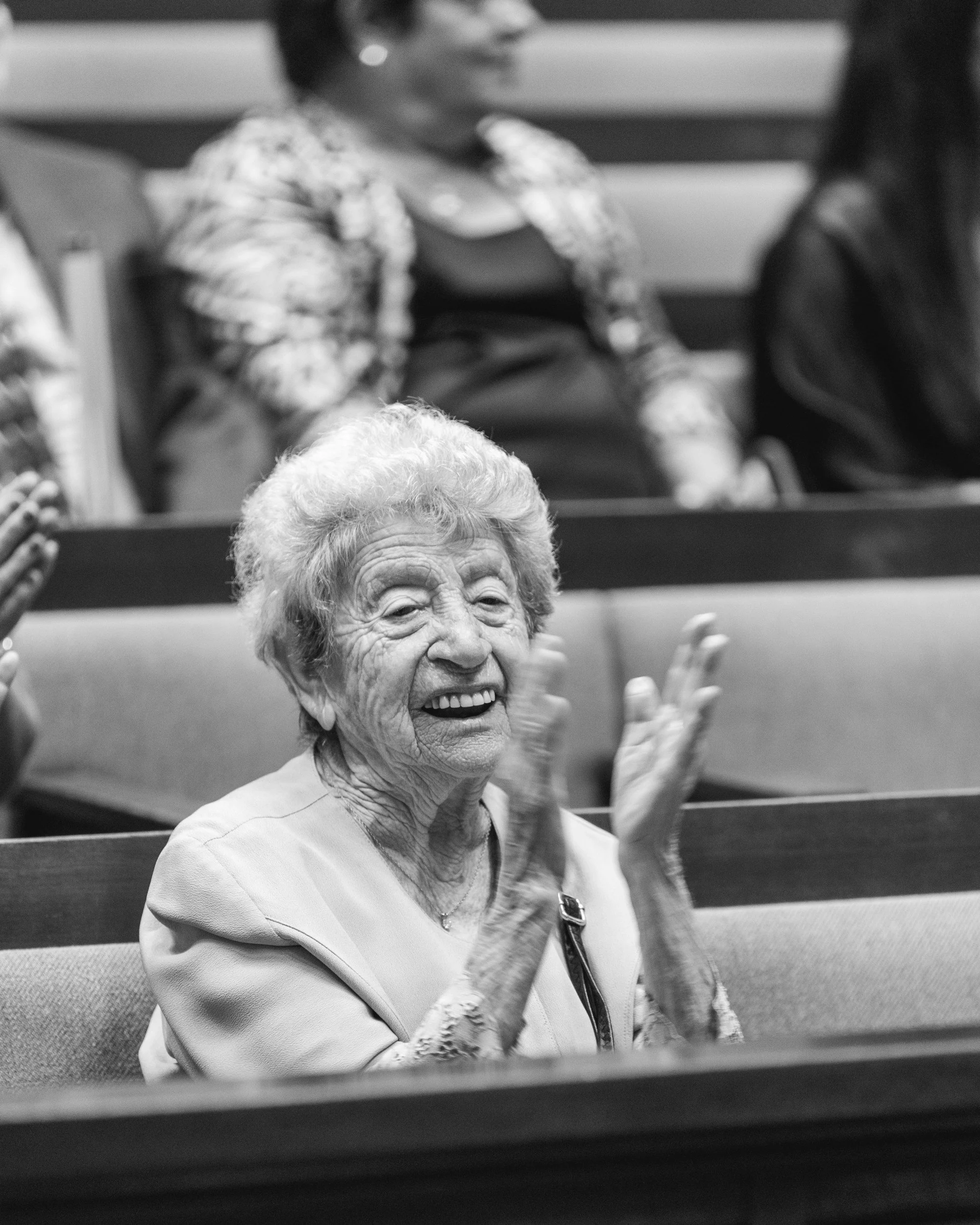 An elderly woman with curly hair claps and smiles in a room with others, who are visible in the background.