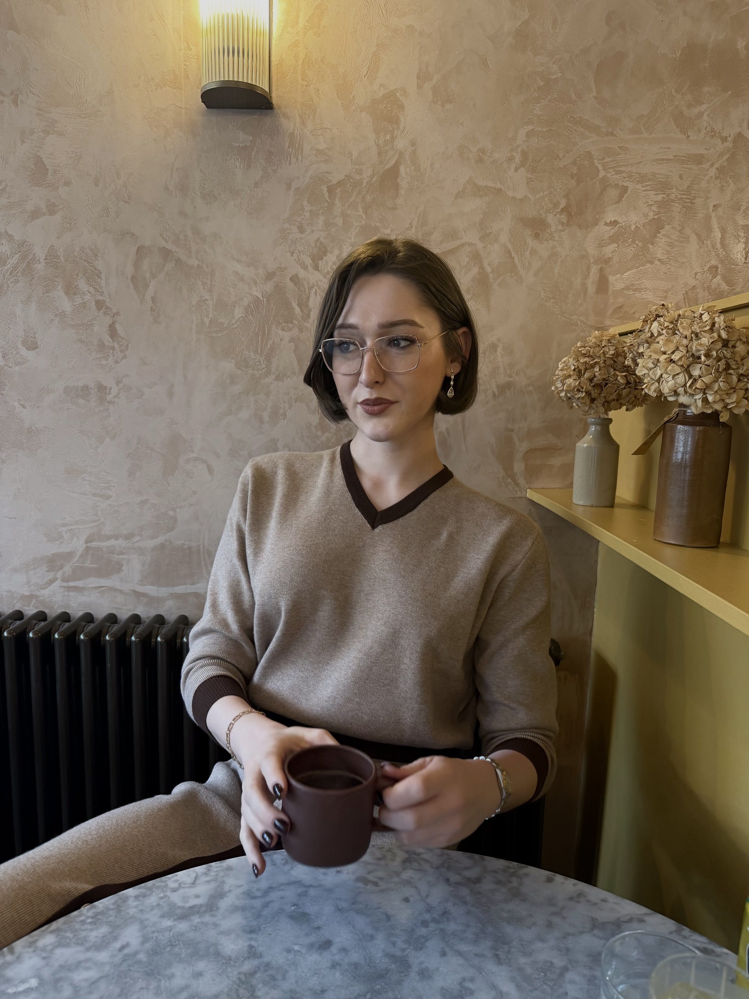 A woman with short brown hair and glasses sitting at a marble table in a cozy, warmly lit room. She is holding a dark brown mug and wearing a matching beige sweater and pants. There are dried hydrangea flowers in vases on a yellow shelf behind her, and a textured beige wall with a wall sconce above.