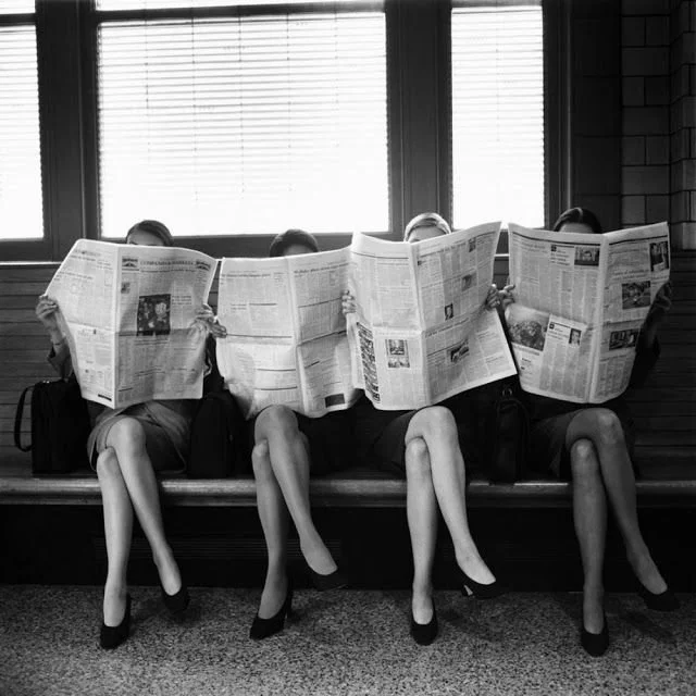 Four women in professional attire sitting on a bench, reading newspapers, with their faces covered by the newspapers, in front of a window with blinds.