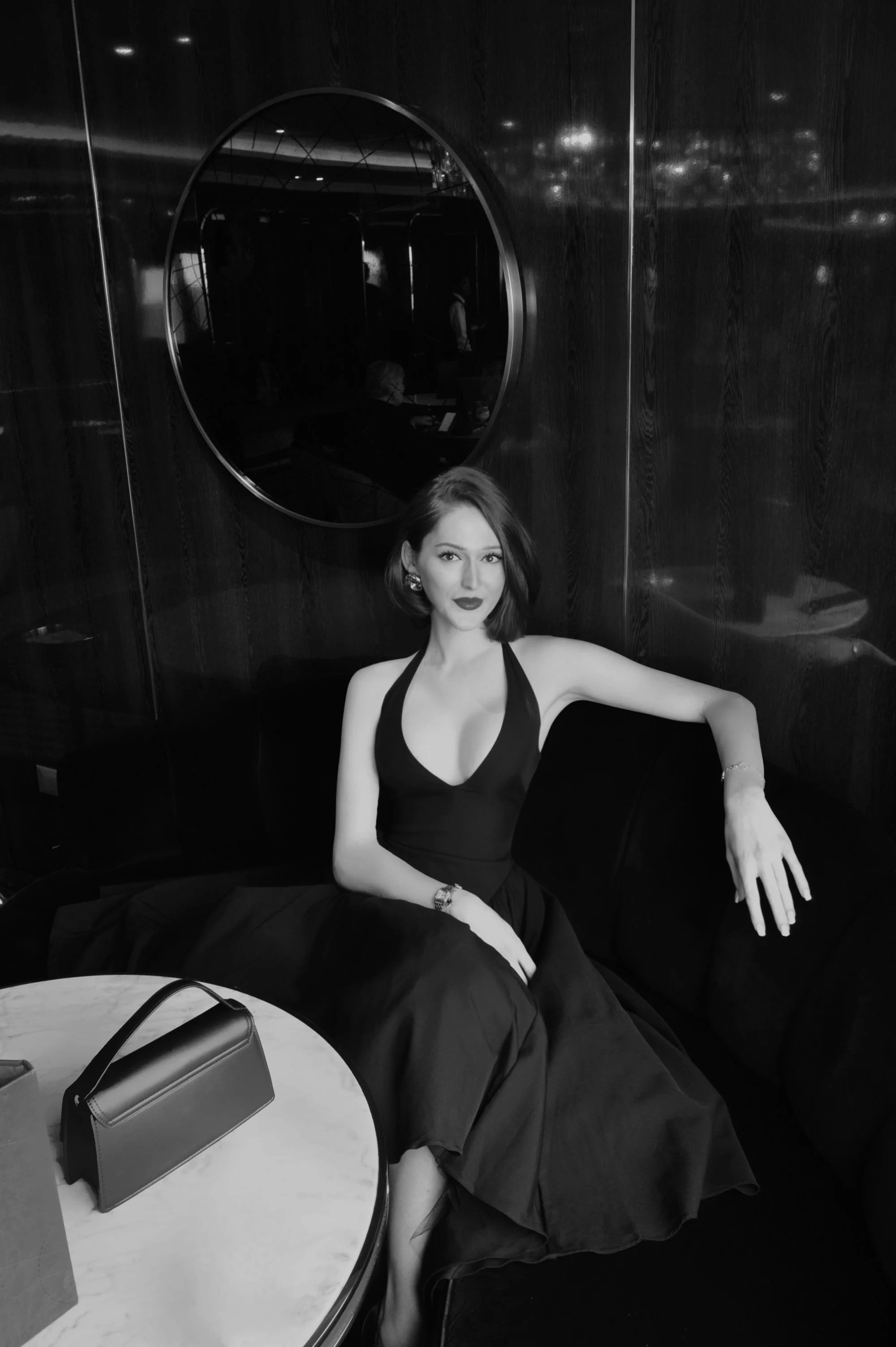 A woman in a black dress sitting on a black velvet sofa in a dimly lit room with dark wooden walls. There is a round marble table in front of her with a black handbag on it. A circular mirror is on the wall behind her, reflecting some figures and lights.