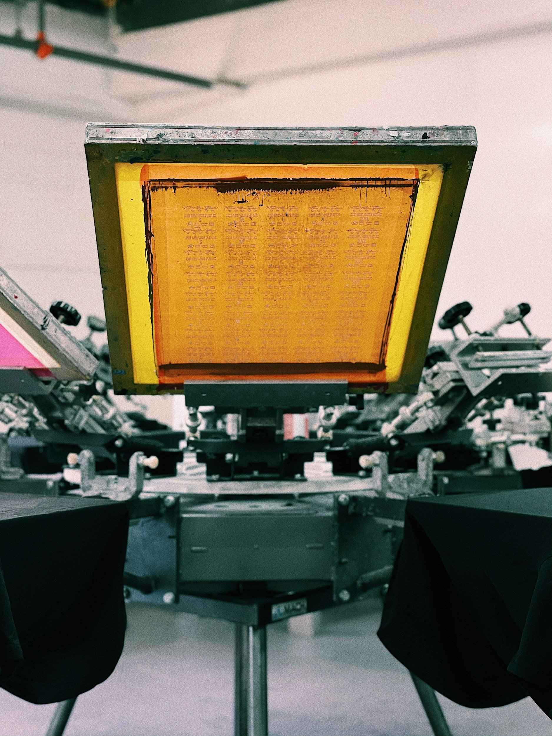 Close-up of a printing screen with orange ink, part of a silk-screen printing machine, in a workshop or studio setting.