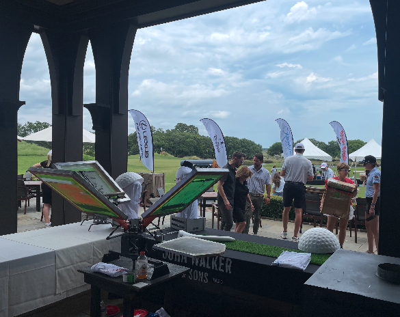 A golf event snack bar with two warming trays, a condiment bottle, and a green-covered display table, overlooking a grassy golf course with people, flags, and tents in the background under partly cloudy skies.
