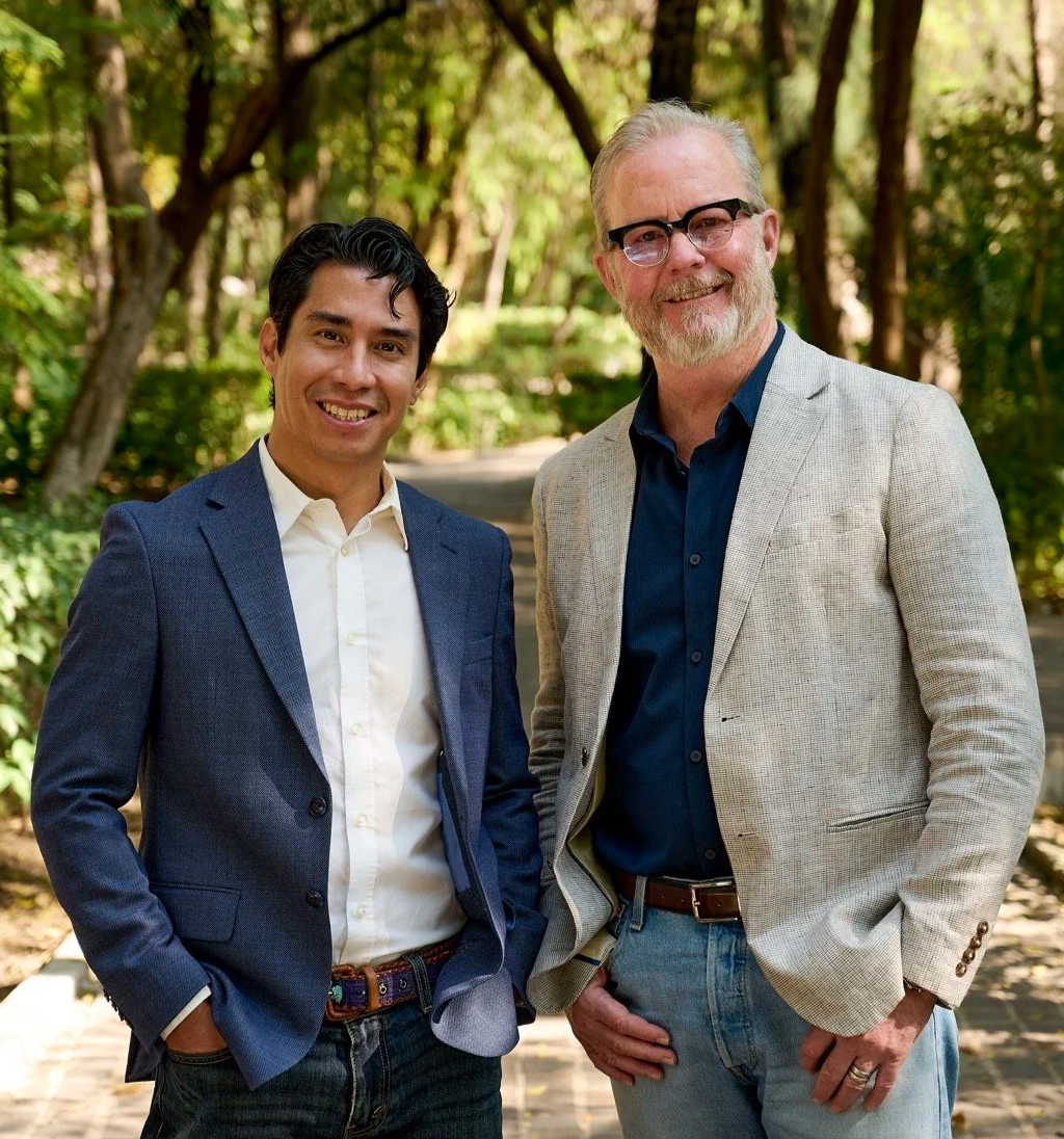 Two men smiling outdoors in a wooded area. One man has black hair and is wearing a blue blazer with a white shirt, and the other has gray hair and a beard, wearing glasses, a light-colored blazer, and a dark shirt.