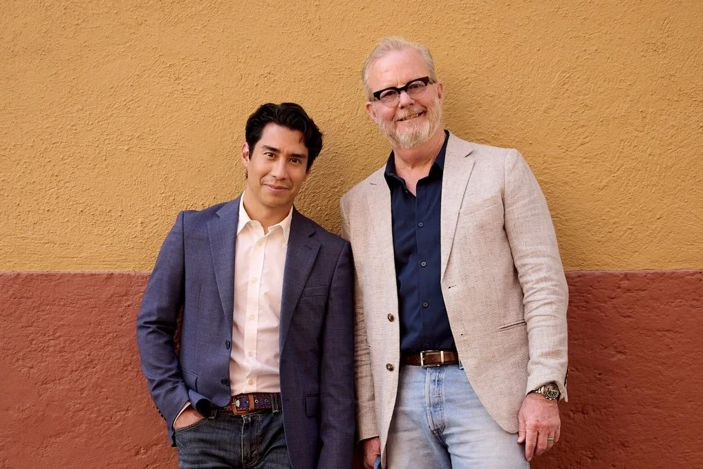 Two men standing against a two-tone yellow and reddish-brown wall, smiling at the camera. The man on the left has dark hair and is wearing a navy blazer over a white shirt, while the man on the right has gray hair, glasses, and is dressed in a light-colored blazer over a navy shirt.