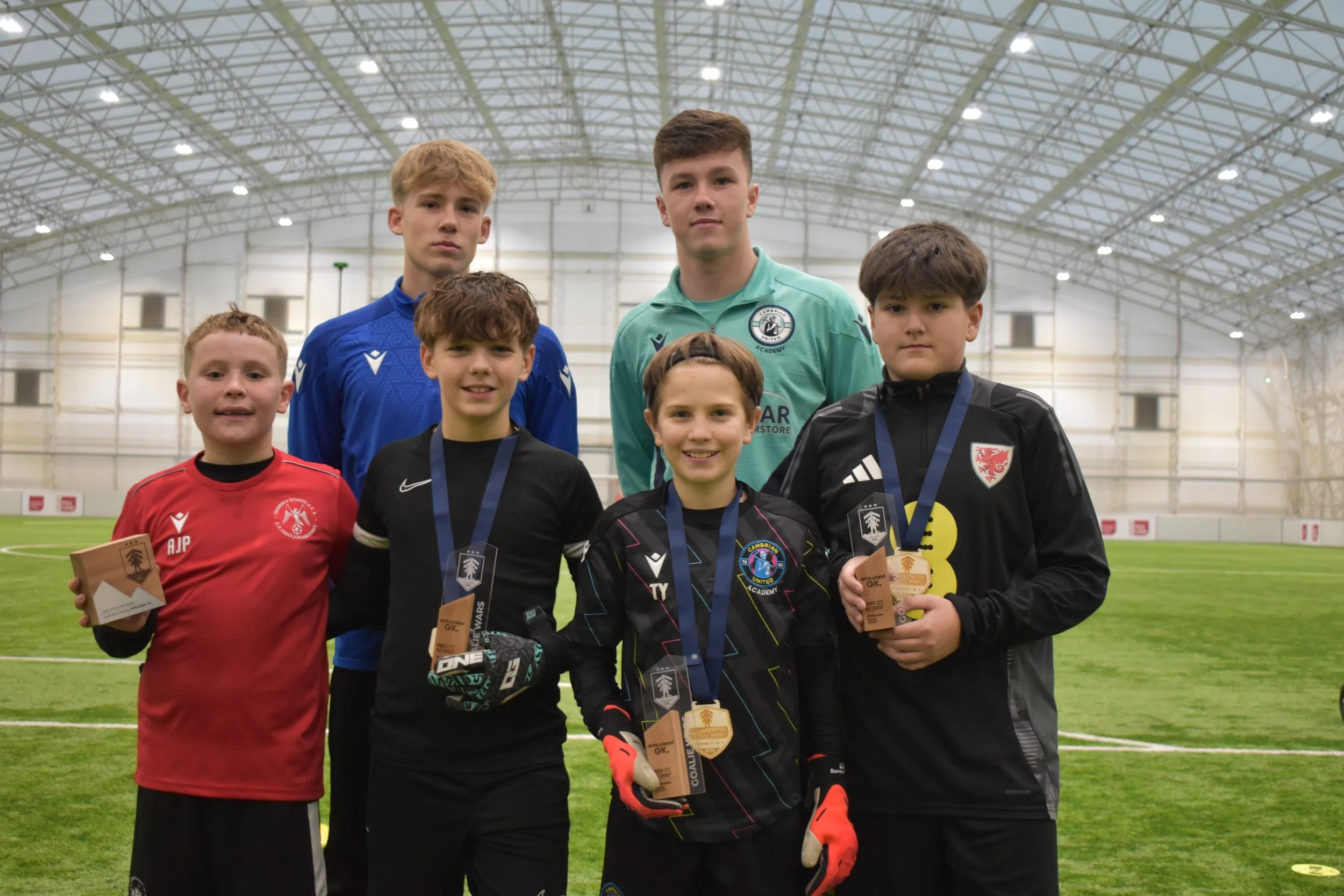 Age group Winner (Under 11, 12 and 13) from the Intelligent GK Goalie Wars on the 20th December 2025.Group of six young boys in soccer uniforms standing inside a large indoor sports facility with a dome-shaped ceiling, some holding awards and medals.