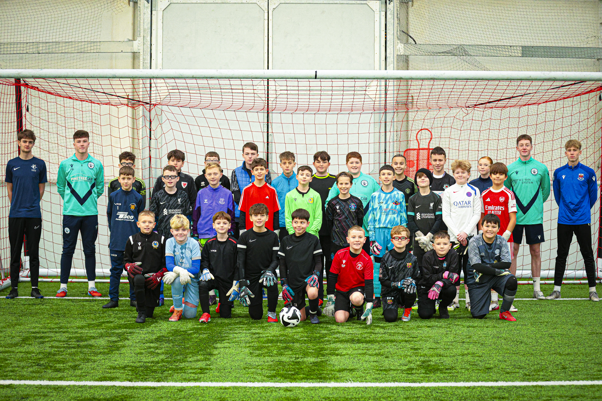 Group photo from Intelligent GK Goalie Wars 20th December 2025 - Group of young boys and teenage players in soccer uniforms standing and kneeling in front of a soccer goal on an indoor field.