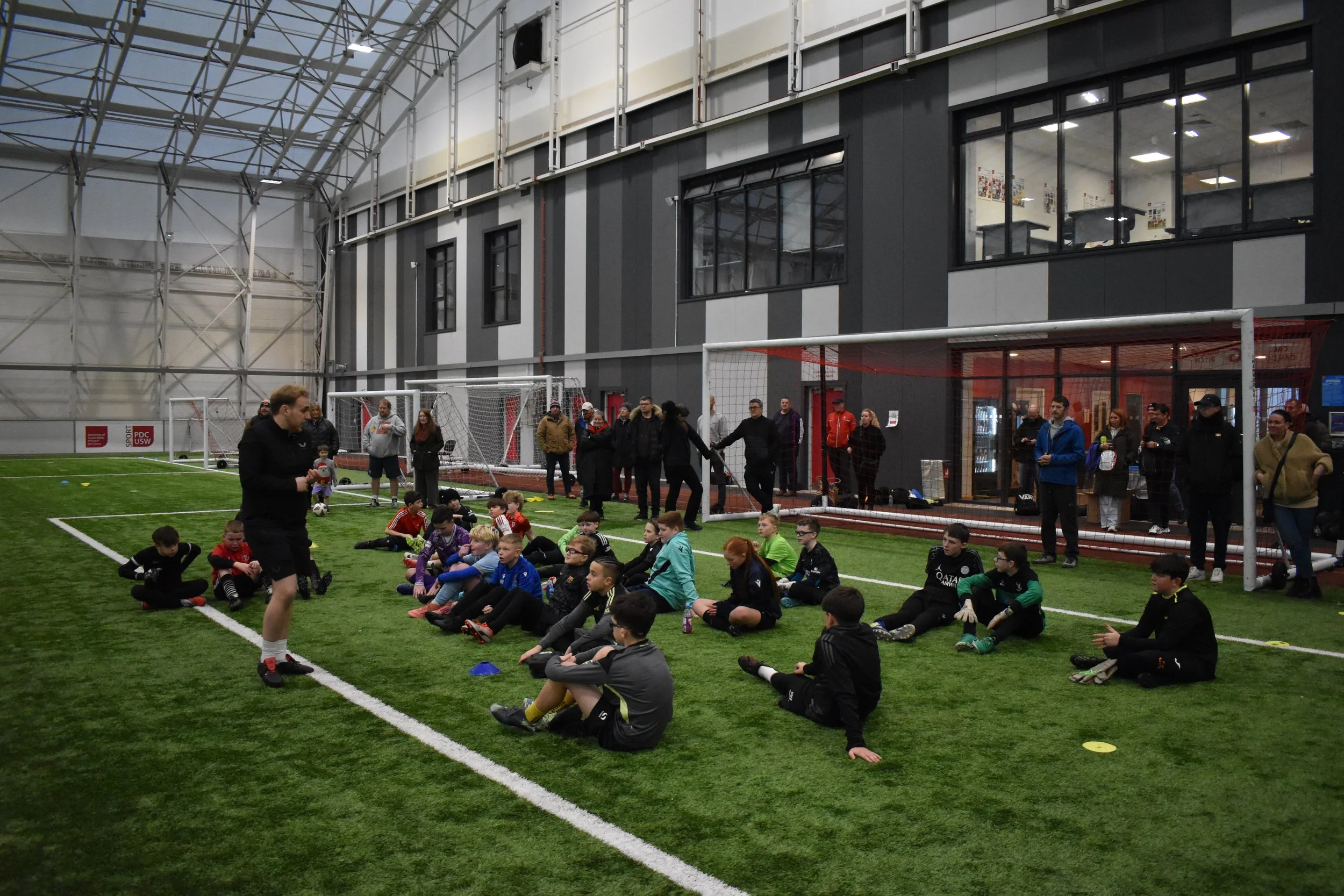 Goalkeeper Wars Briefing - Children participating in a soccer training session inside an indoor sports facility, sitting on the artificial turf, while a coach speaks to them. Spectators and parents are watching from behind a glass wall.