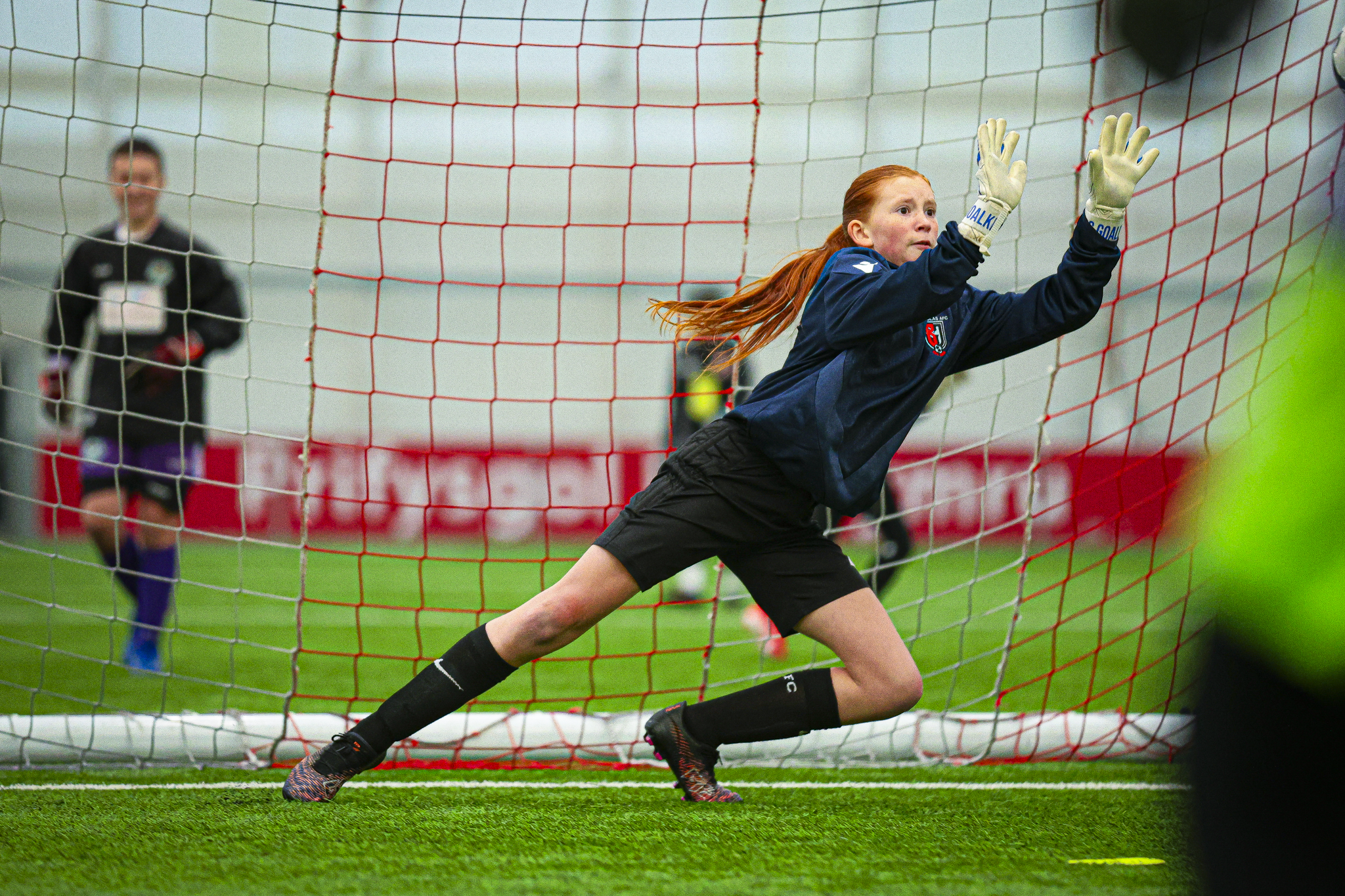 A female soccer goalkeeper with long red hair, wearing a navy blue jersey, black shorts, and gloves, diving to her right to block a shot near the goal, with a blurred background of an indoor soccer field.