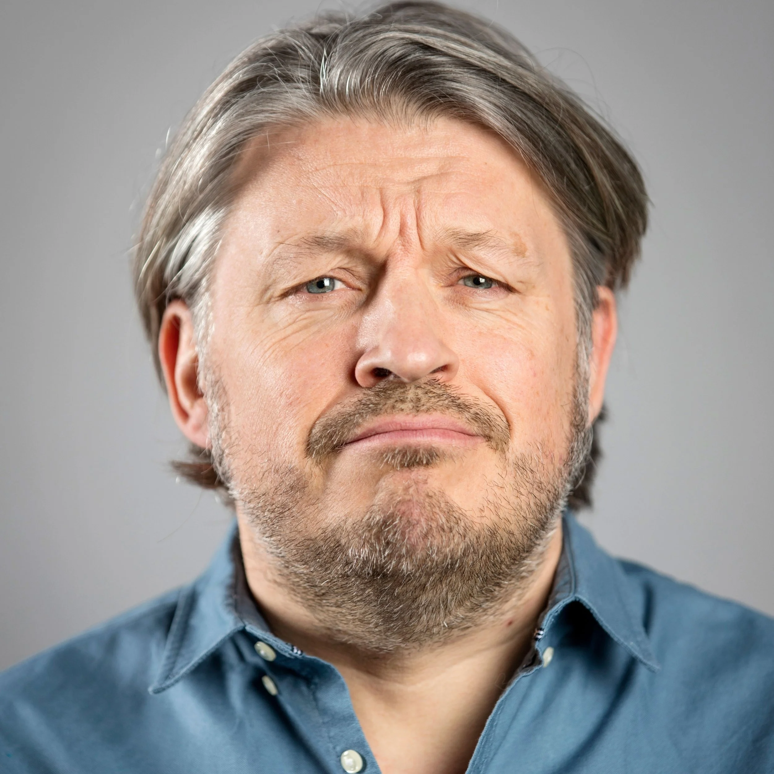 Close-up of a middle-aged man with gray hair, blue eyes, and a beard, wearing a blue collared shirt, making a skeptical or confused facial expression.
