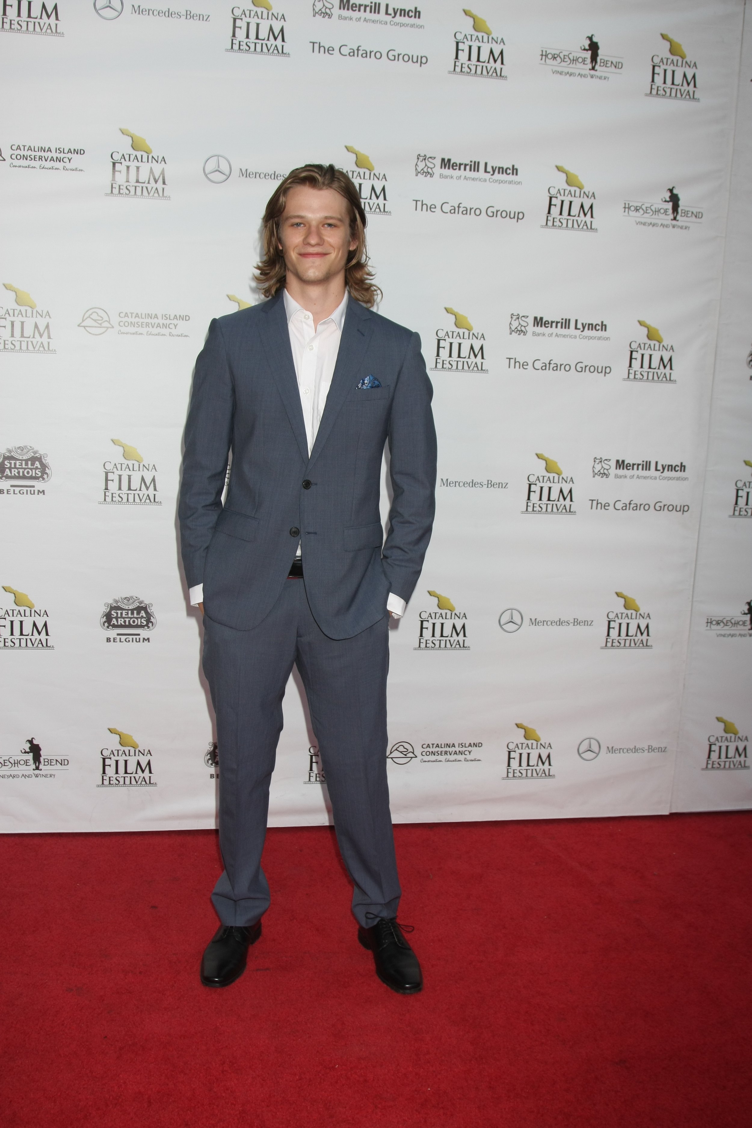 Young man in a gray suit with a white shirt, standing on a red carpet at a film festival, with a backdrop featuring various sponsor logos and the event's name.