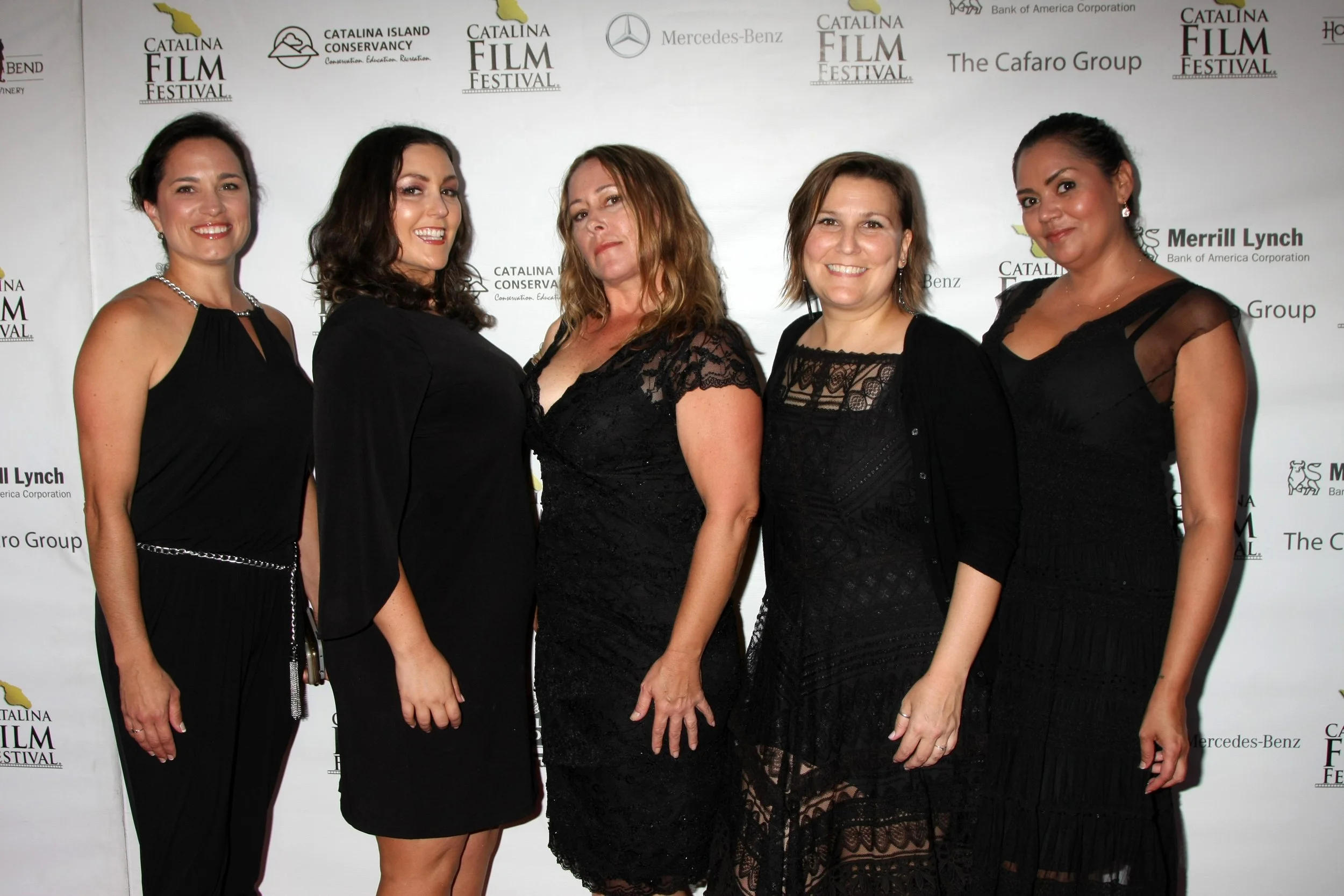 Five women dressed in black at a film festival event, standing in front of a white backdrop with logos, smiling and posing for the camera.