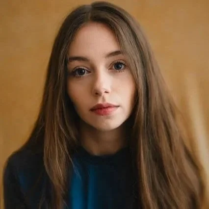 Close-up of a young woman with blue eyes, long brown hair, and a neutral expression, wearing a beige top against a dark background.