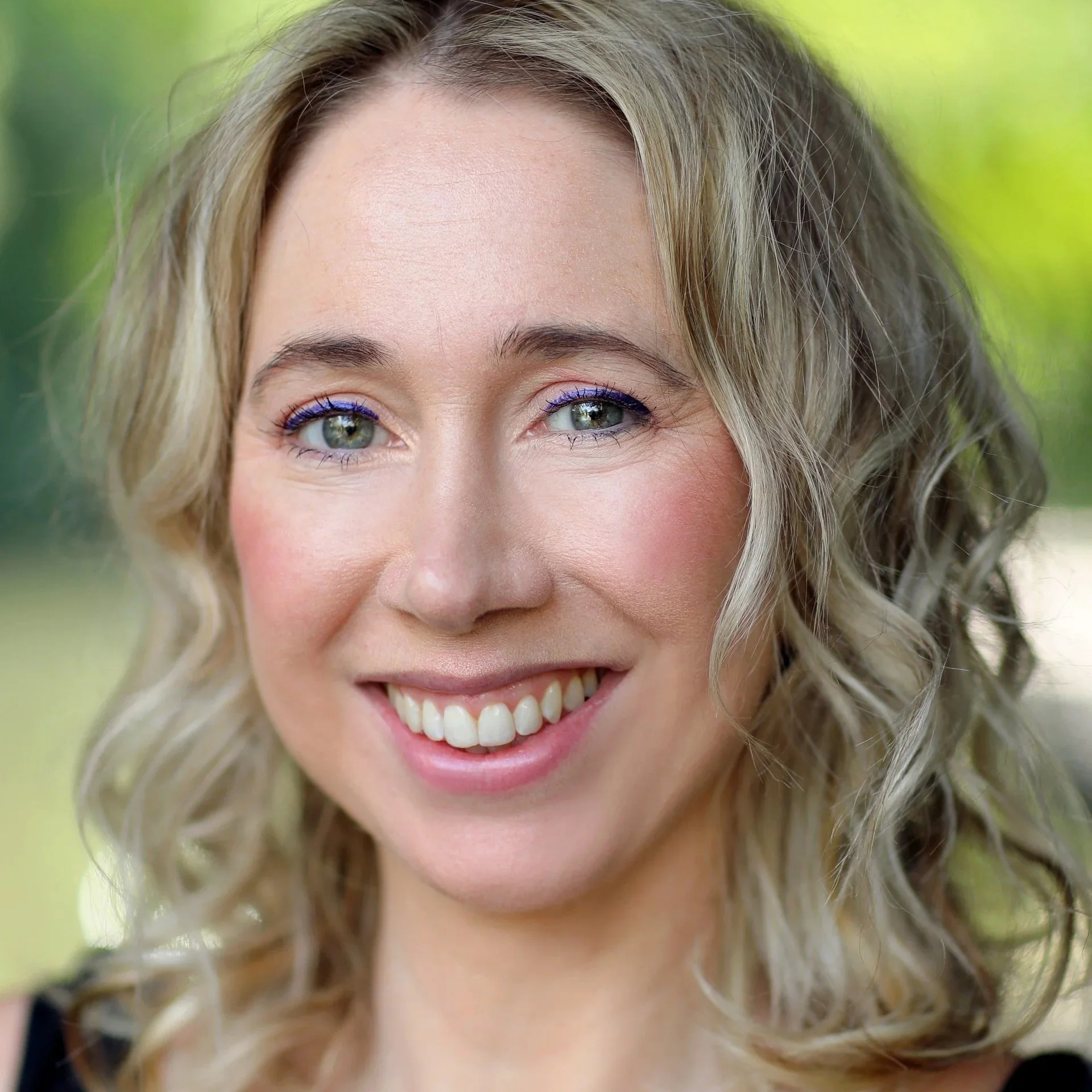 A woman with light blonde curly hair, blue eyes, and a bright smile posing outdoors with a blurred green background.
