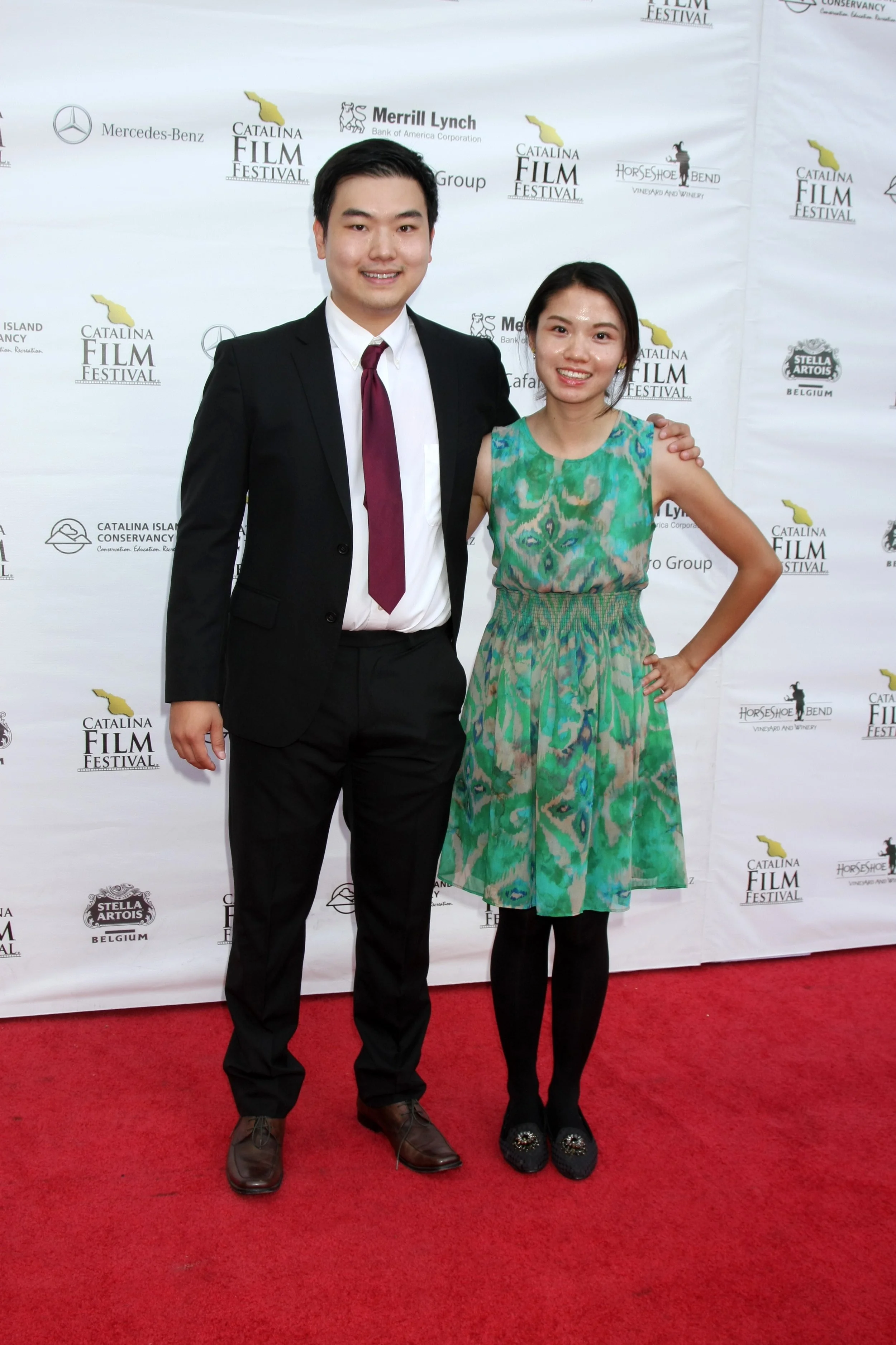A young man in a black suit with a white shirt and maroon tie standing next to a young woman in a colorful green and blue dress on a red carpet at the Catalina Film Festival. They are smiling and posing for the photo with a step and repeat banner in 