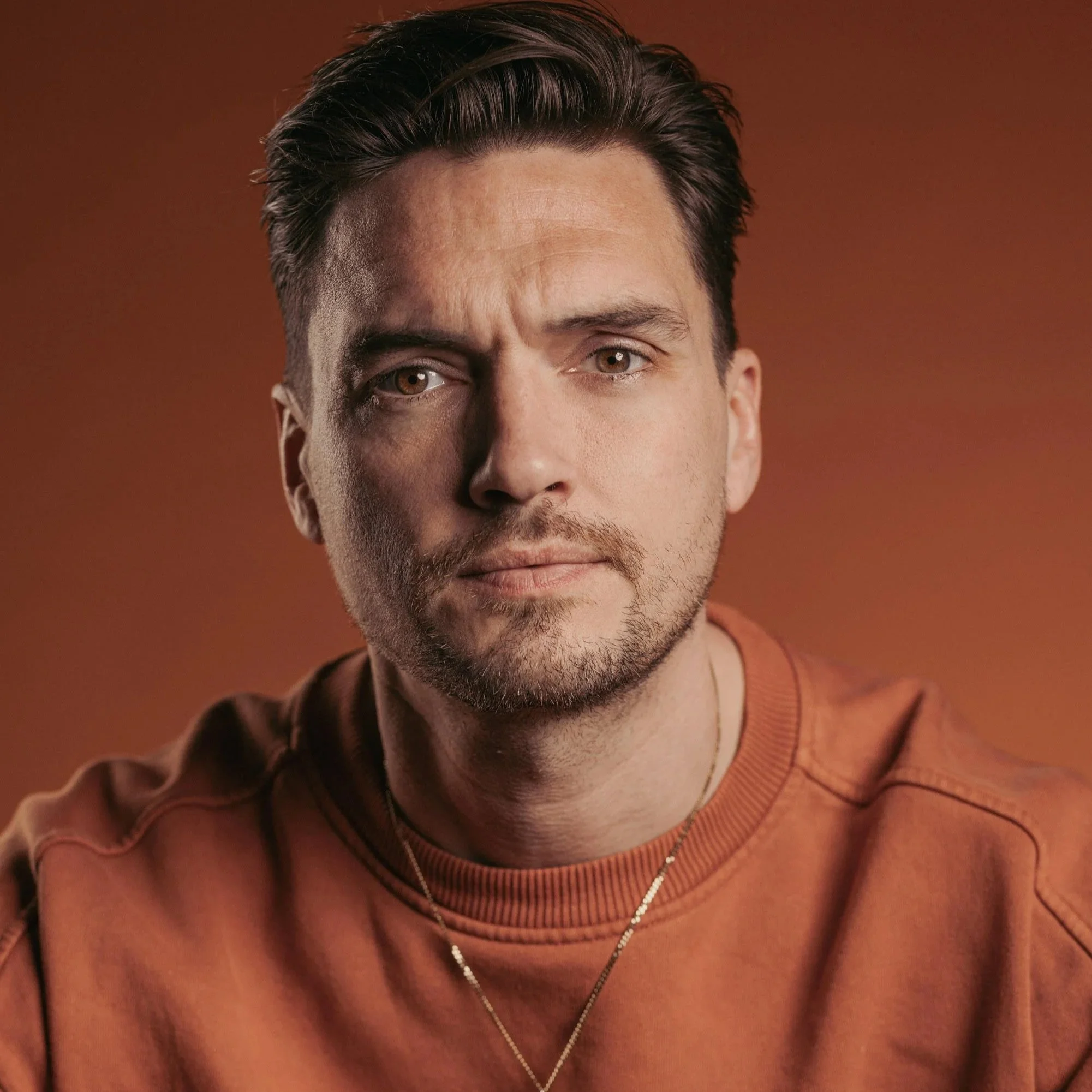 Close-up of a man with short dark hair, a beard, and brown eyes, looking directly at the camera with a neutral expression.