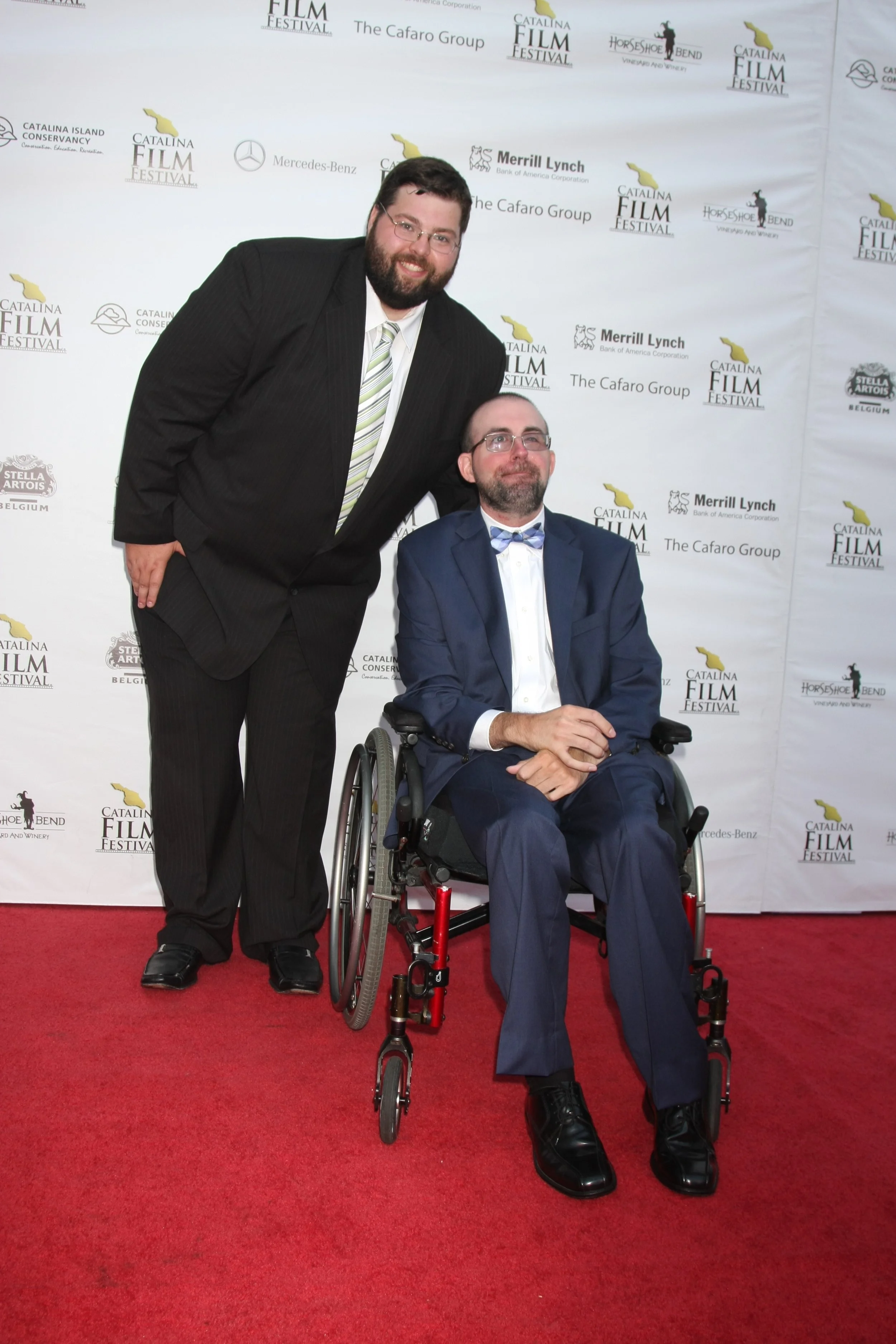 Two men at an event, one standing with arm on a seated man's shoulder. The seated man is in a wheelchair, wearing a blue suit and bow tie. The standing man is in a dark suit with a striped tie. The backdrop is decorated with various sponsor logos and