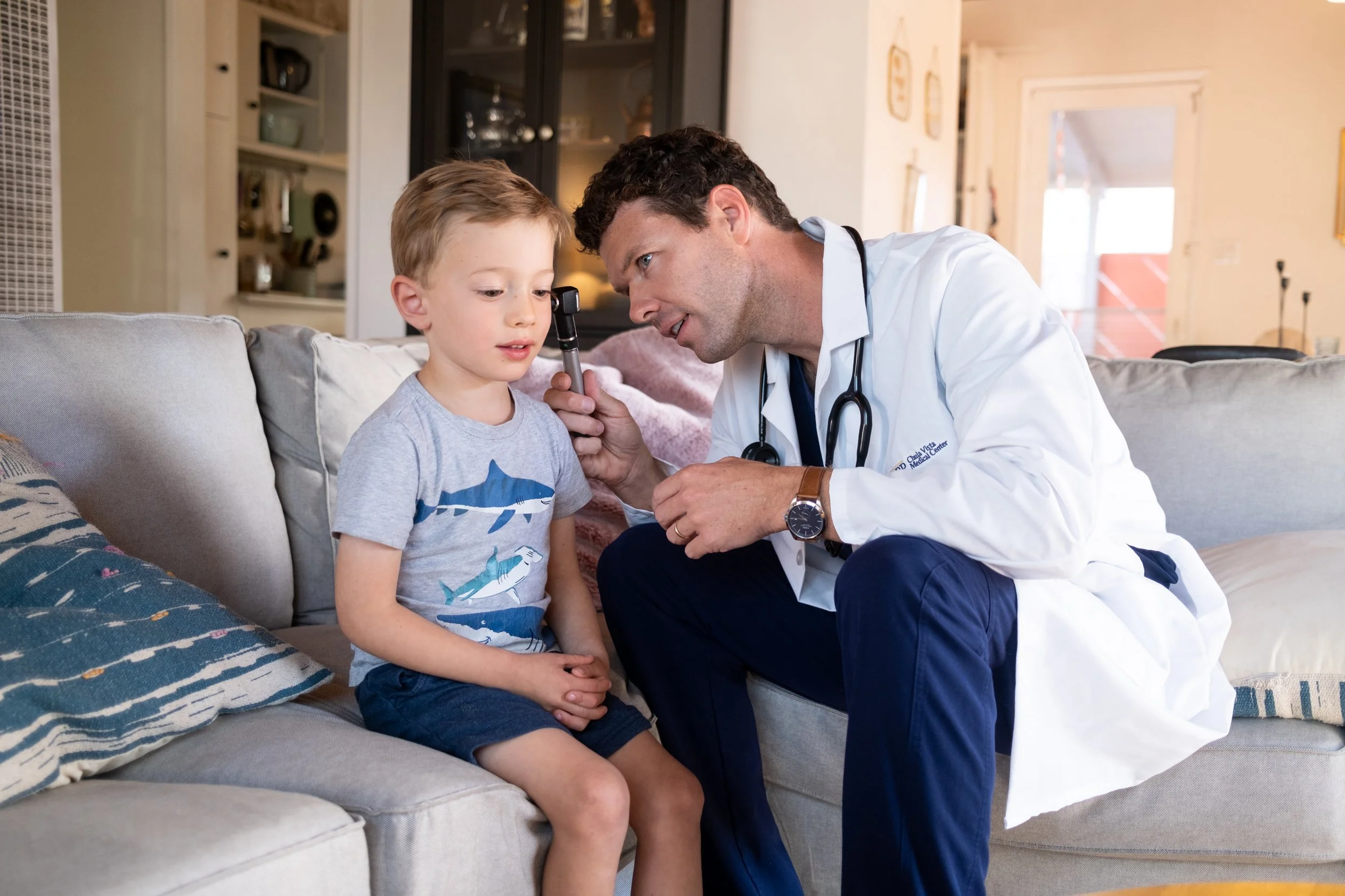 A doctor examining a young boy's ear with an otoscope in a living room.
