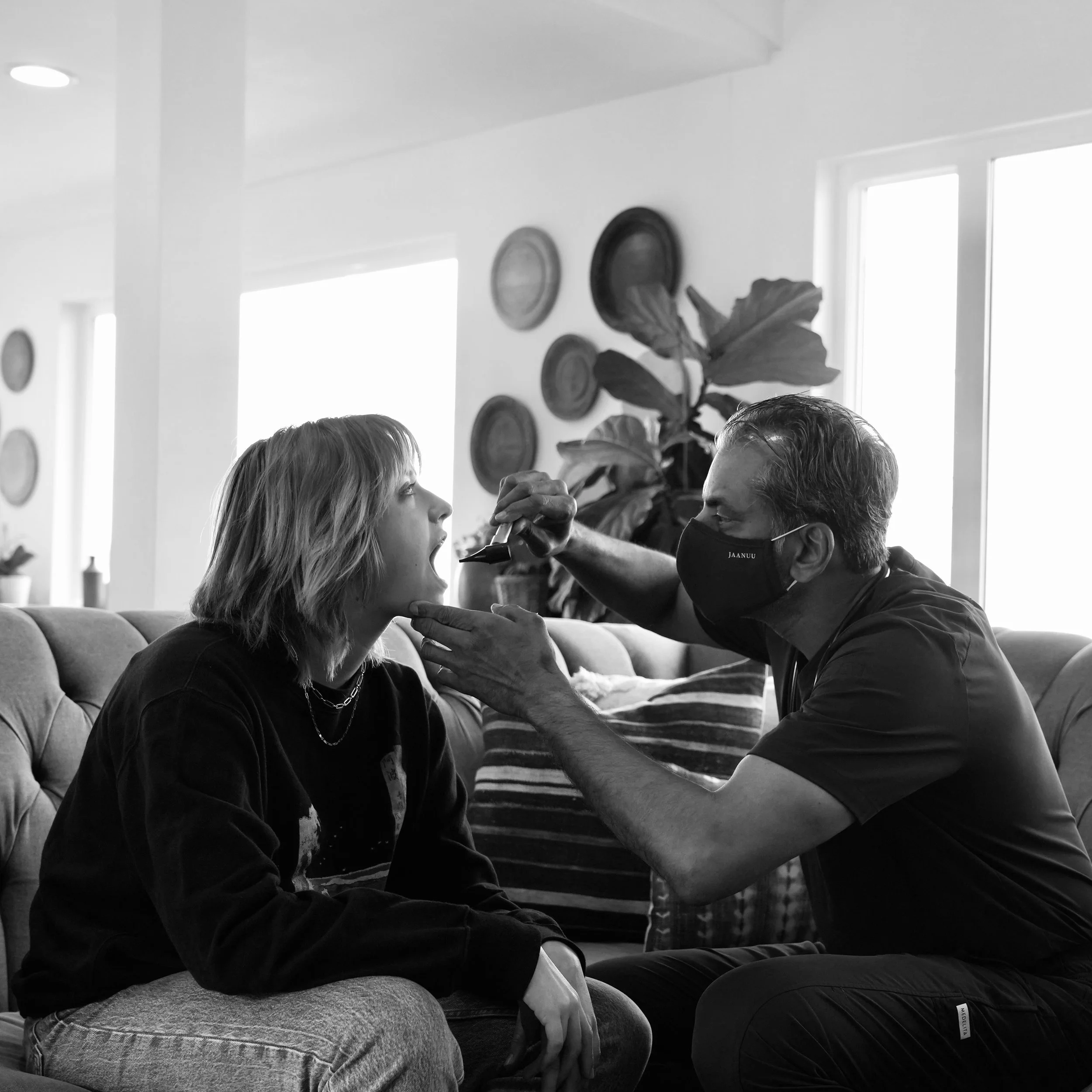 A woman sitting on a couch while a man in a face mask administers a nasal swab test in her nose inside a well-lit living room with wall decorations and a large plant.