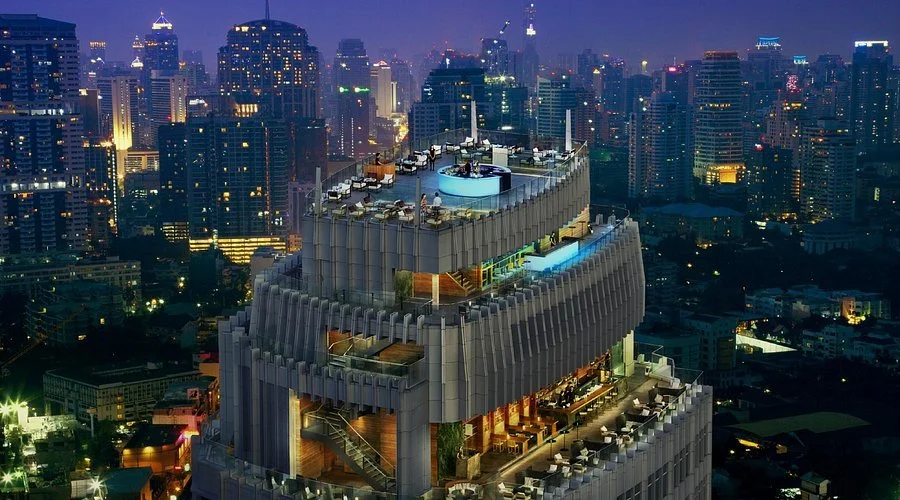 Night view of a tall skyscraper with multiple outdoor terraces and a rooftop pool, overlooking a city skyline filled with illuminated buildings.