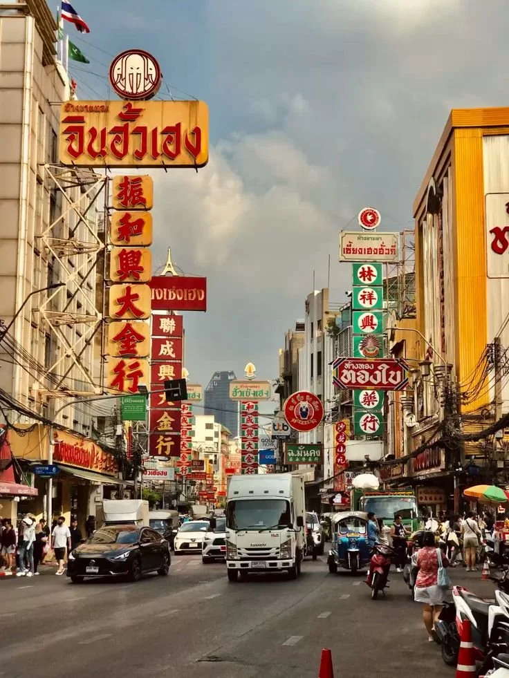 A busy street scene in an Asian city with various signs in Thai and Chinese characters, pedestrians, vehicles, and some storefronts.
