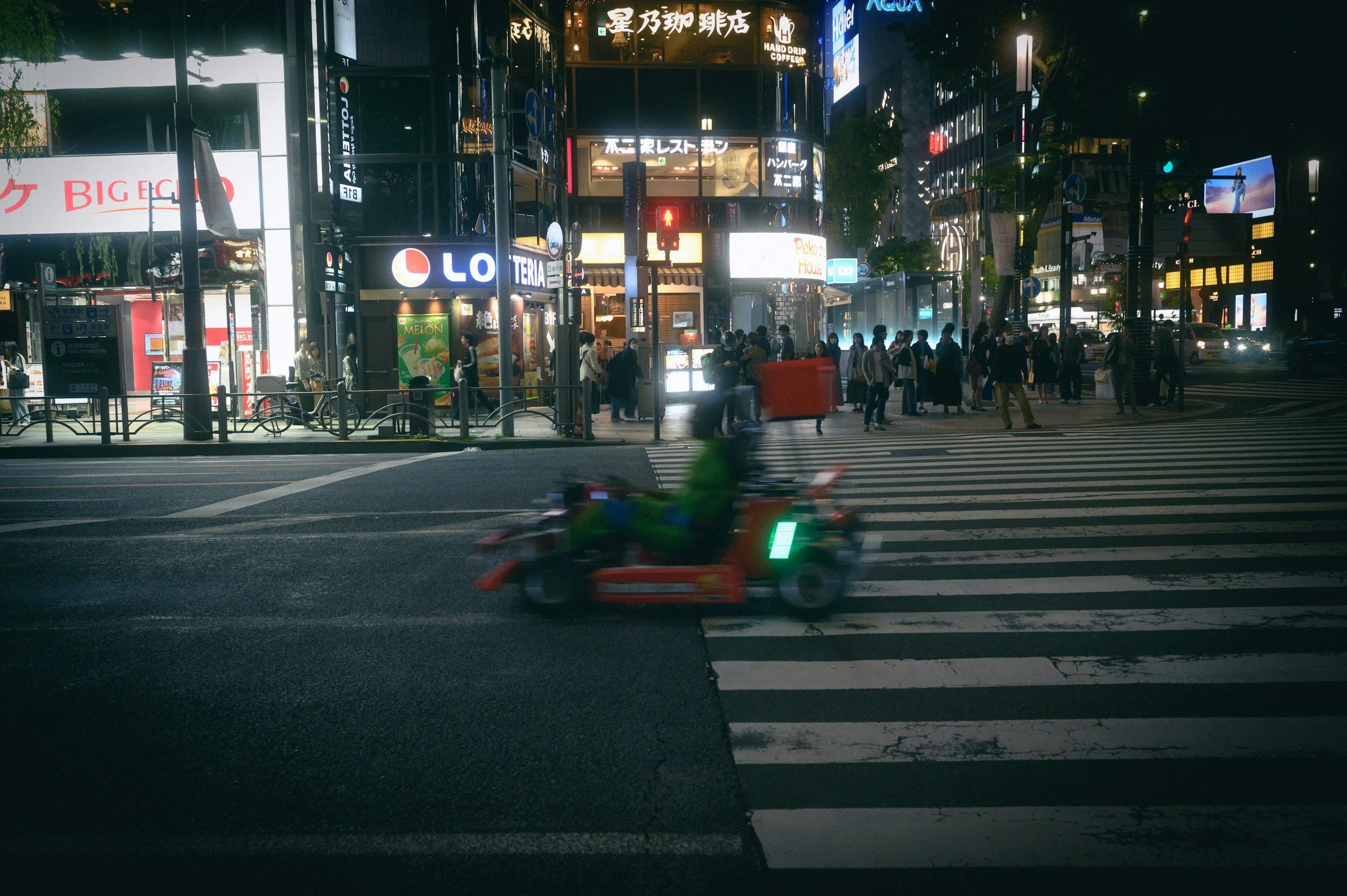Night scene of a busy city crosswalk with pedestrians waiting and a moving bicycle rickshaw in the foreground. Illuminated store signs and billboards line the background.