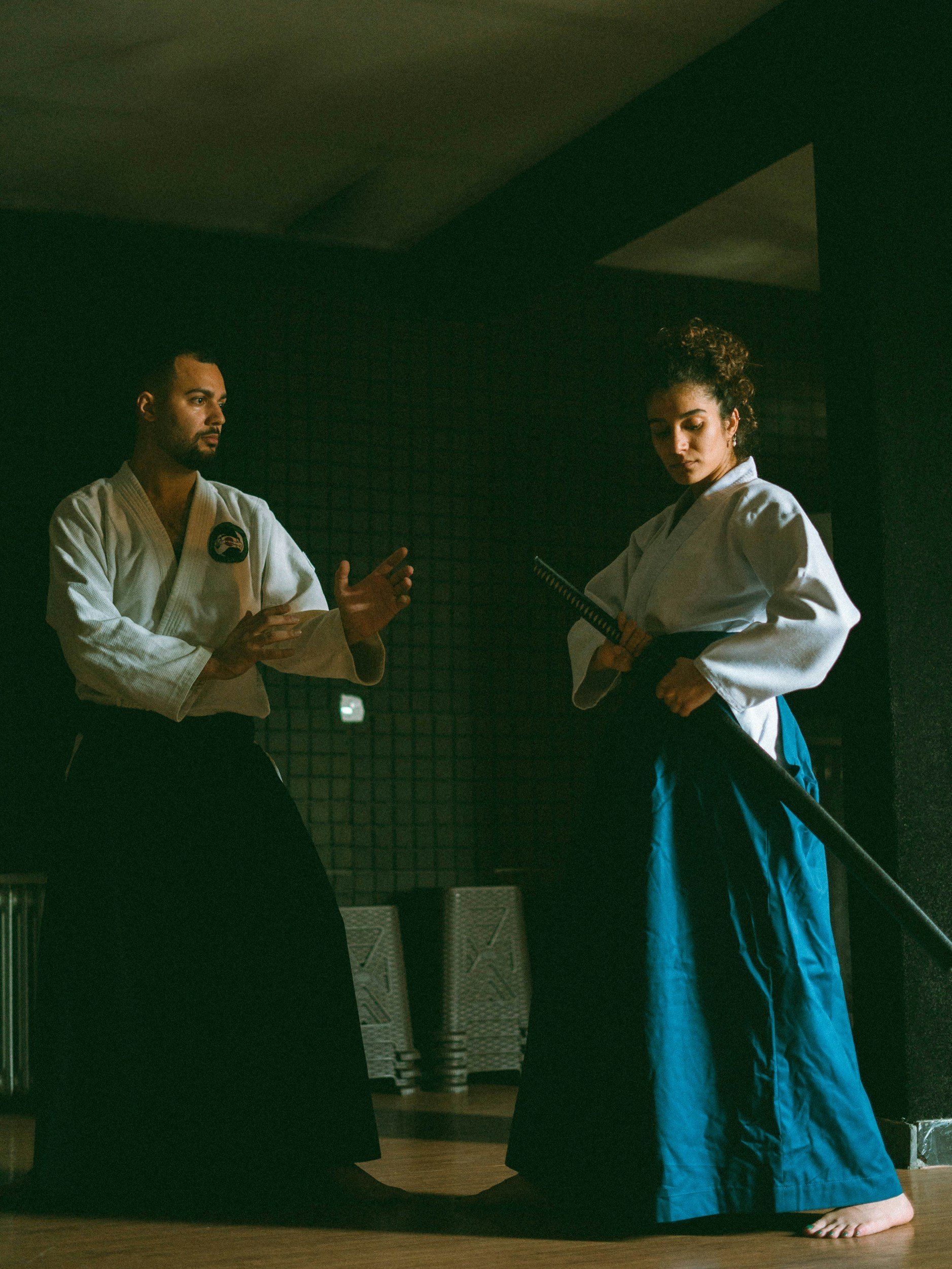Two martial artists in traditional uniforms practicing with a sword and a smaller weapon in a dimly lit dojo.