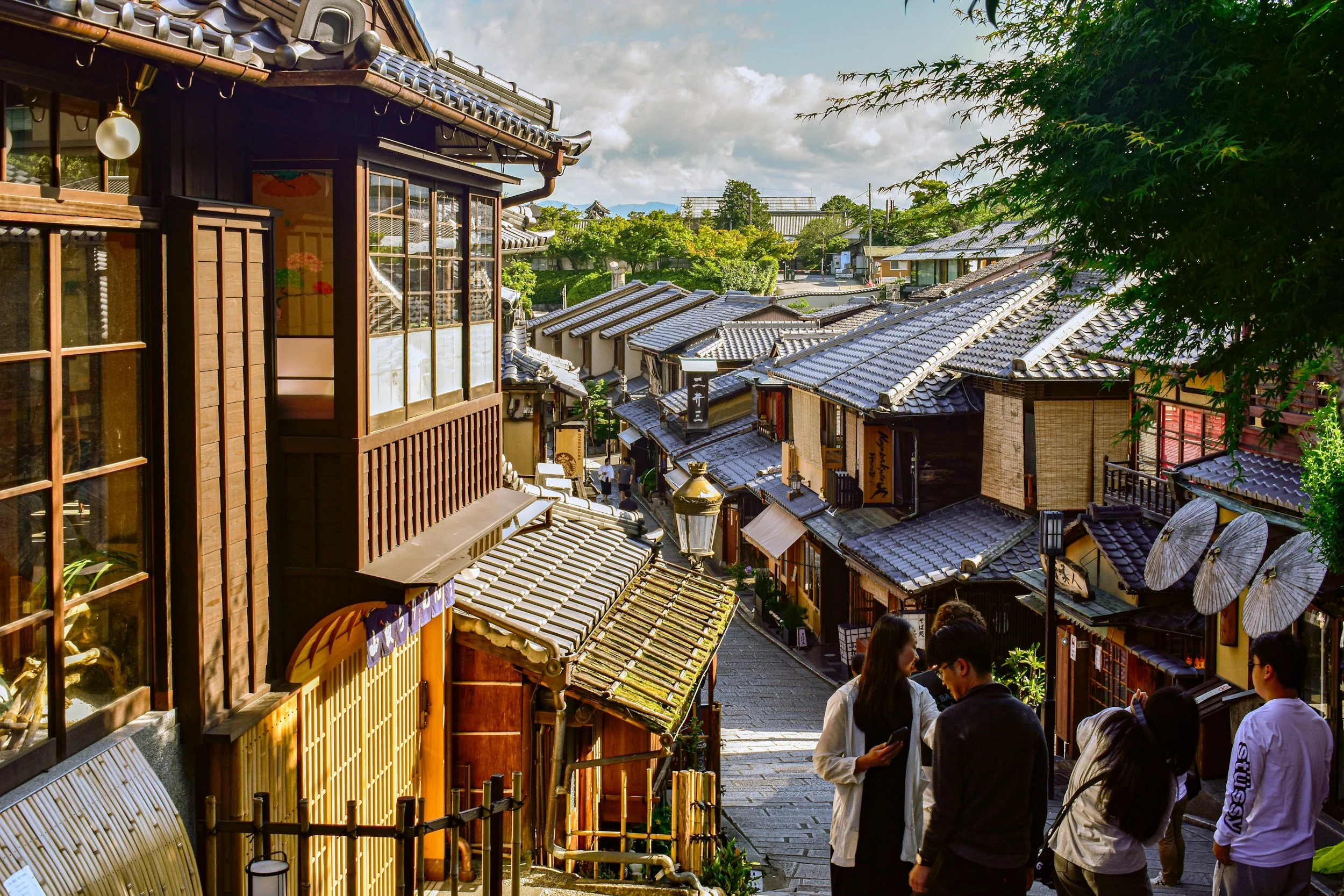 Street view of traditional Japanese buildings with tiled roofs, some lanterns and umbrellas on the sides, and a group of people chatting at the bottom right, with a partly cloudy sky overhead.