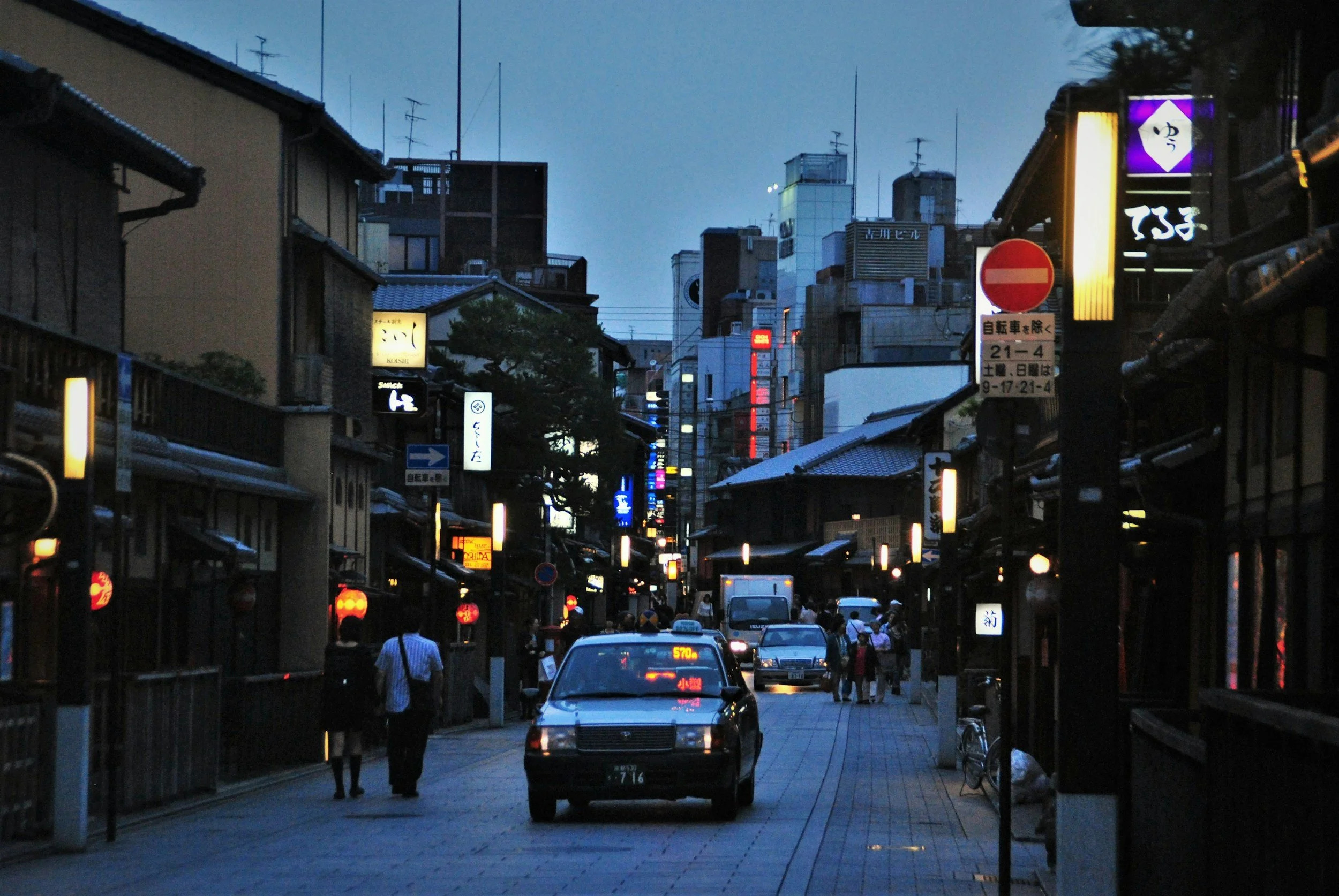 A street scene in Japan during dusk with illuminated signs, pedestrians, and cars, featuring traditional buildings and modern high-rise structures in the background.