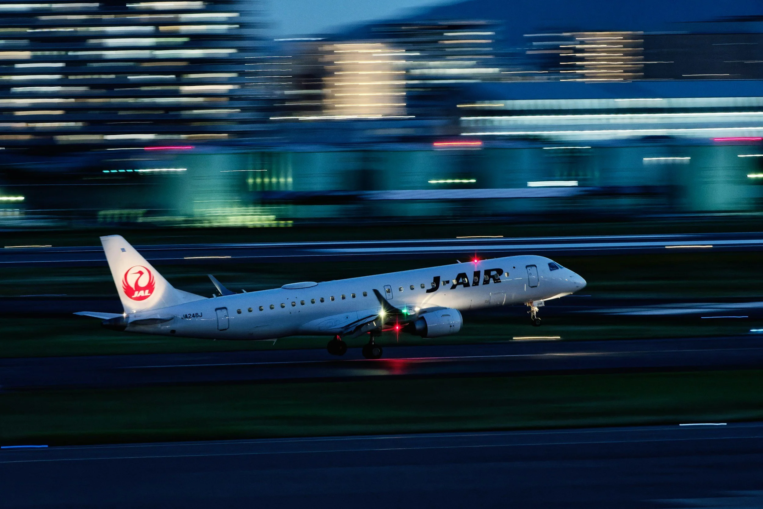 JAL airplane taking off from runway at night with city lights and blurred buildings in the background.