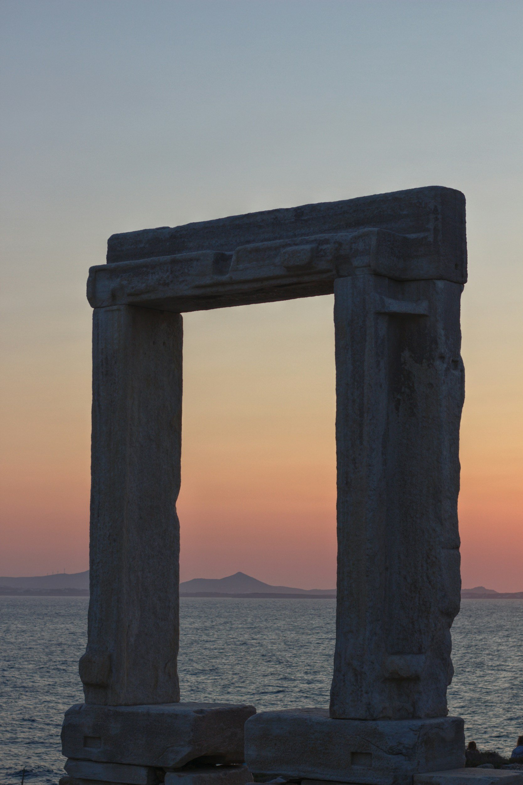 Ancient stone structure with two vertical stones supporting a horizontal stone lintel, set against a sunset sky over the sea with distant islands.