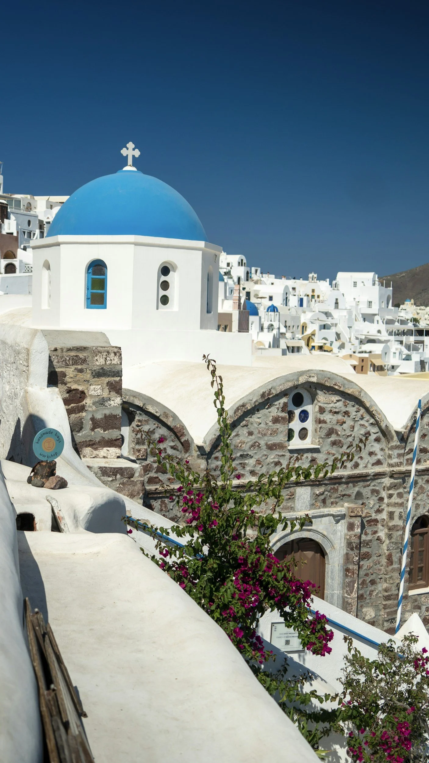 White buildings with blue domes in Santorini, Greece, featuring colorful flowers and clear blue sky.