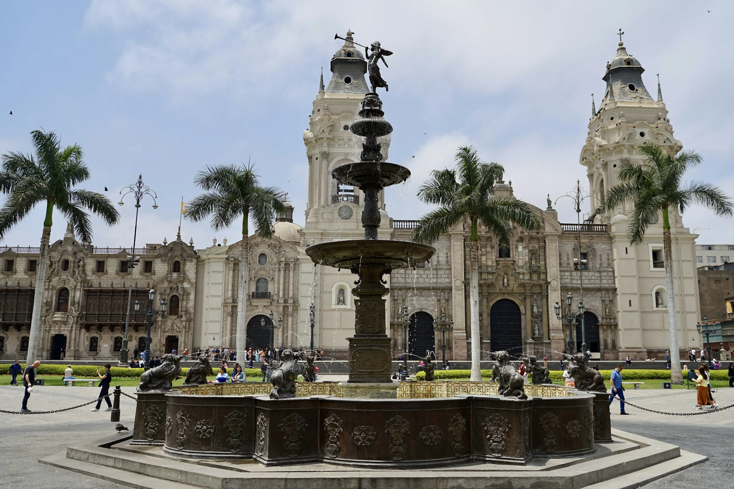 Square in front of a historic church with twin towers, a fountain with a sculpture on top, palm trees, and people walking around.