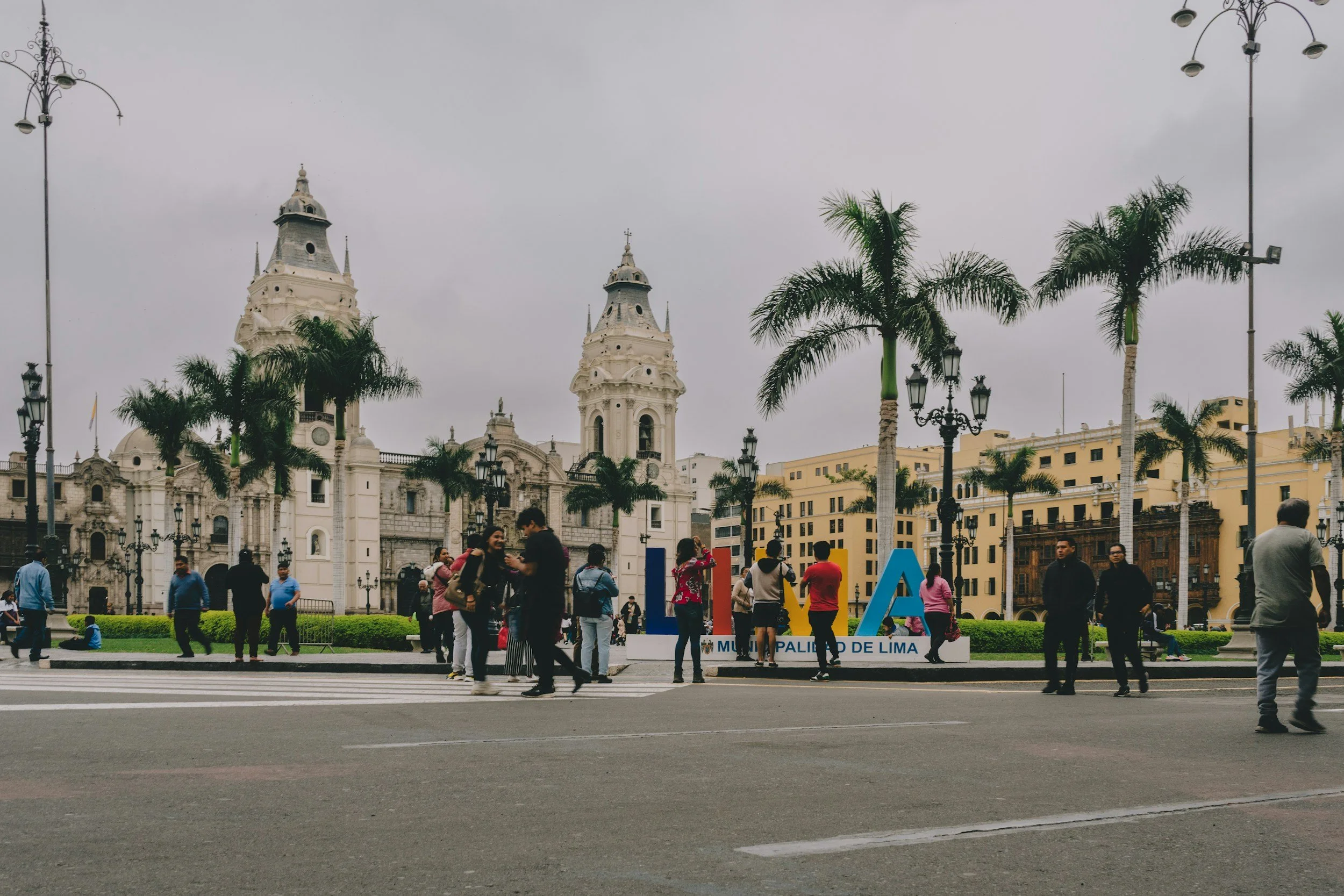 A busy city square in Lima, Peru, with people crossing a crosswalk, in front of a historic white cathedral and palm trees, under an overcast sky.