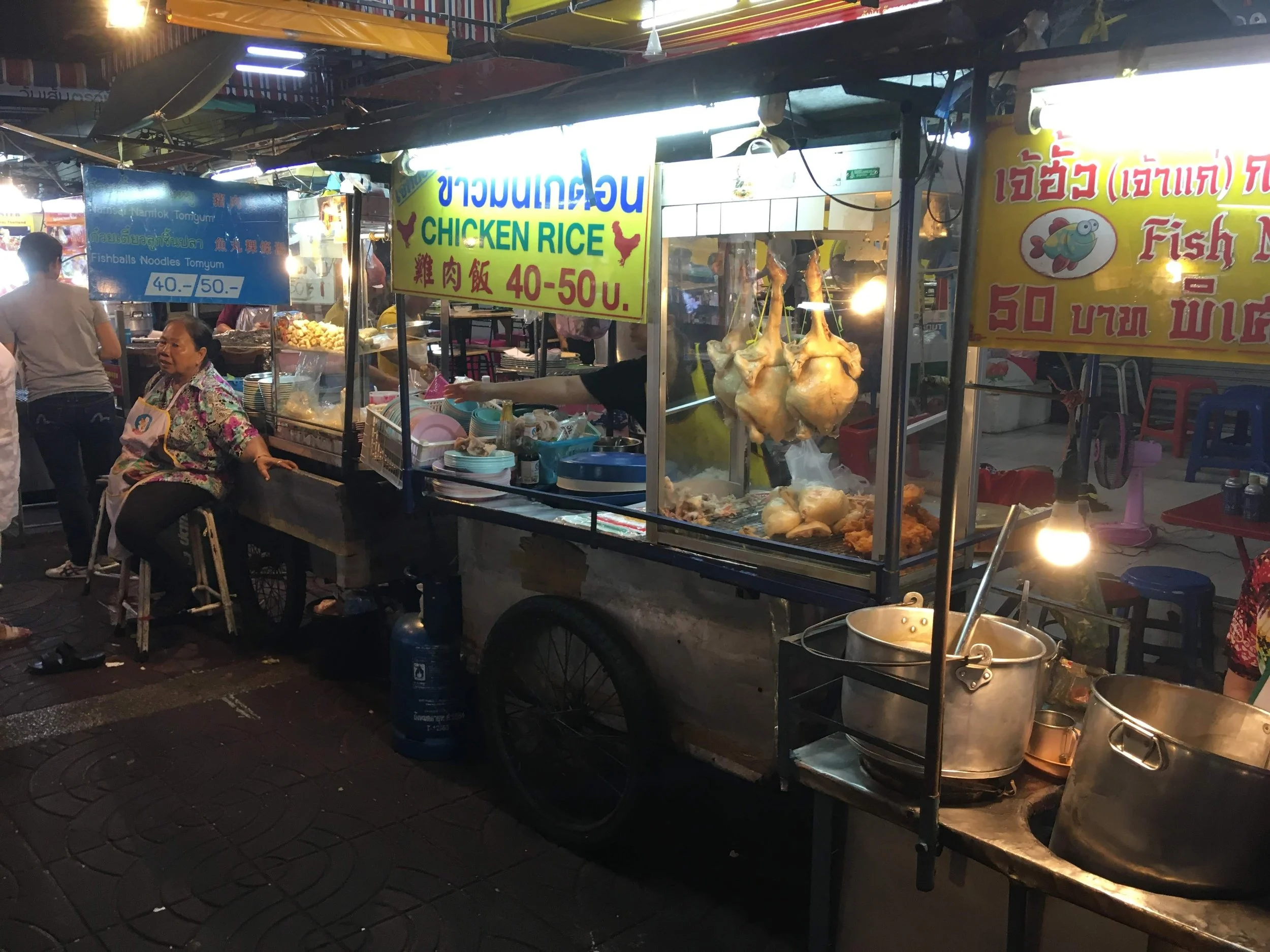 Night market food stall with hanging chicken, fried chicken, and signs in English and Thai, selling chicken rice and fish ball. Customers nearby.