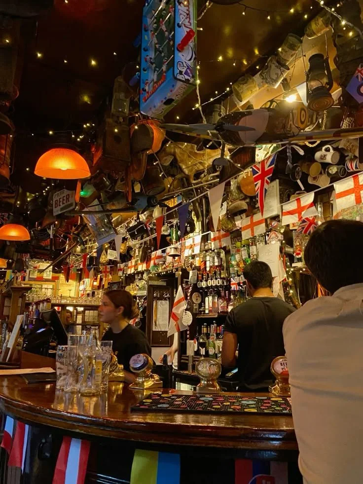 Decorated pub or bar with British flags, string lights, bottles behind the bar, and patrons sitting at the counter.