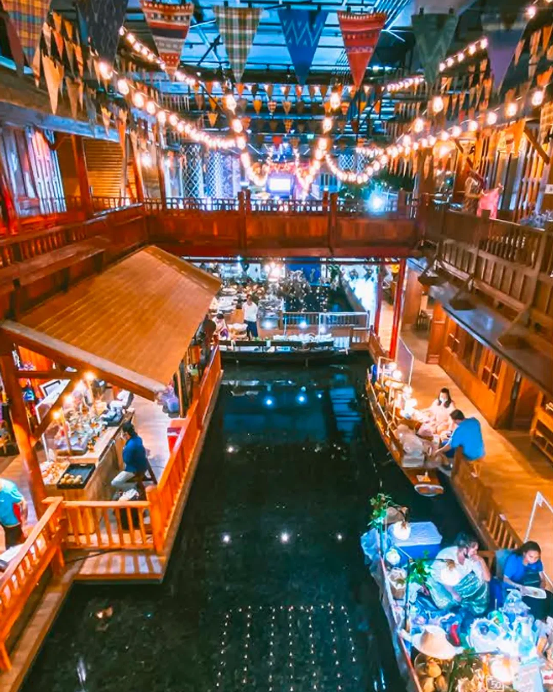 Interior of a two-story wooden restaurant or bar with hanging string lights and colorful triangular banners, overlooking a central pond with people sitting near the water.