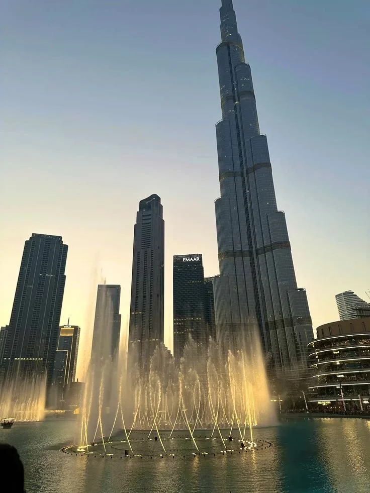 Tall skyscraper with fireworks display in front, city skyline at dusk, fountain with lit water jets in a body of water.