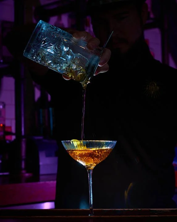 A bartender pouring a drink from a shaker into a cocktail glass on a bar counter, with low lighting and a dark background.