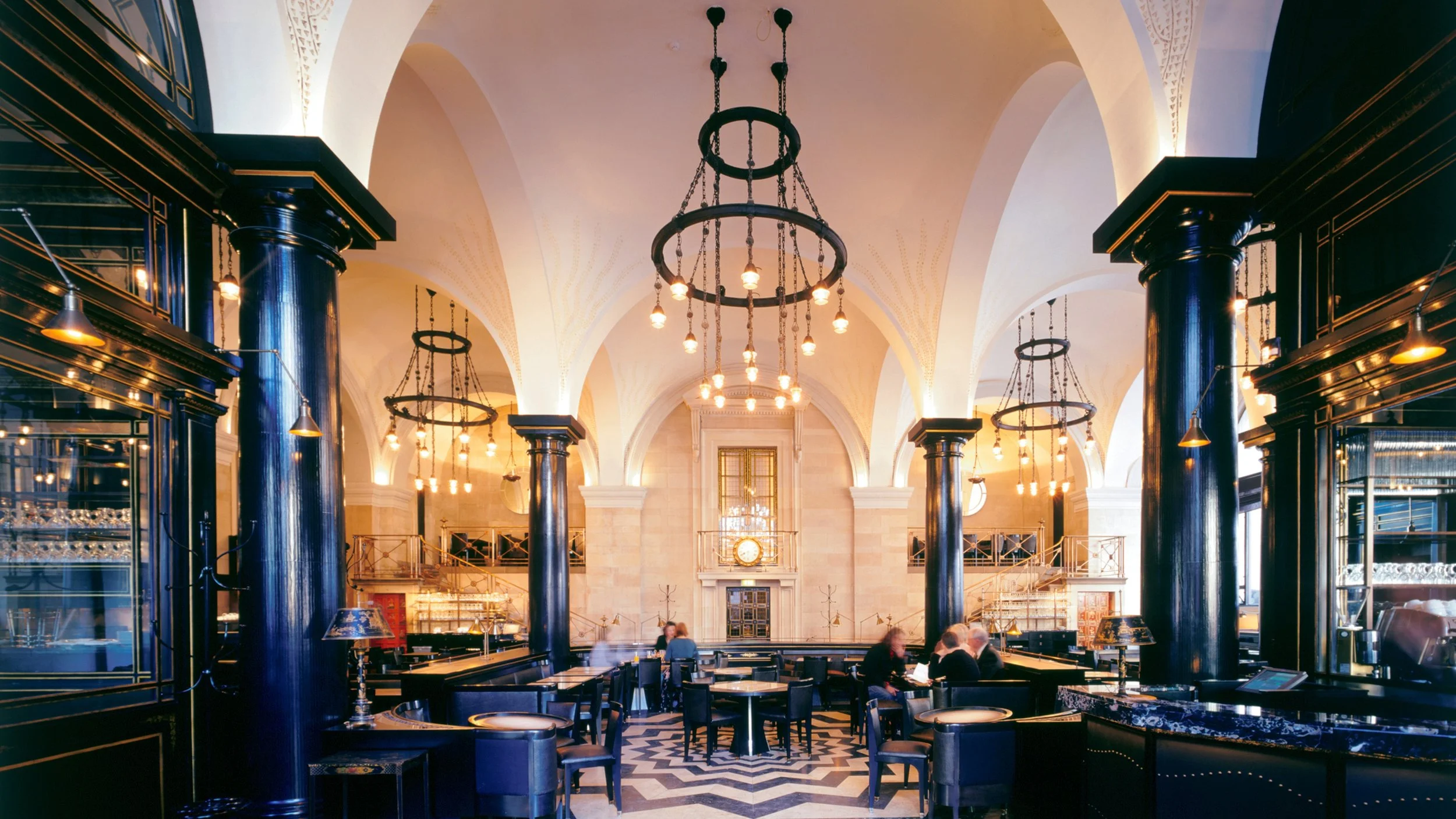 Elegant restaurant interior with high arched ceilings, black columns, chandeliers, and modern decor, with patrons seated at tables.