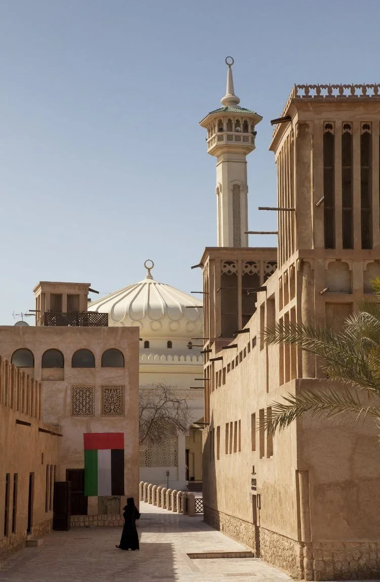 Traditional Middle Eastern architecture with beige buildings, a mosque with a white dome, and a minaret in the background. A woman dressed in black is walking in the shaded alleyway, with an Iraqi flag hanging on a wall.
